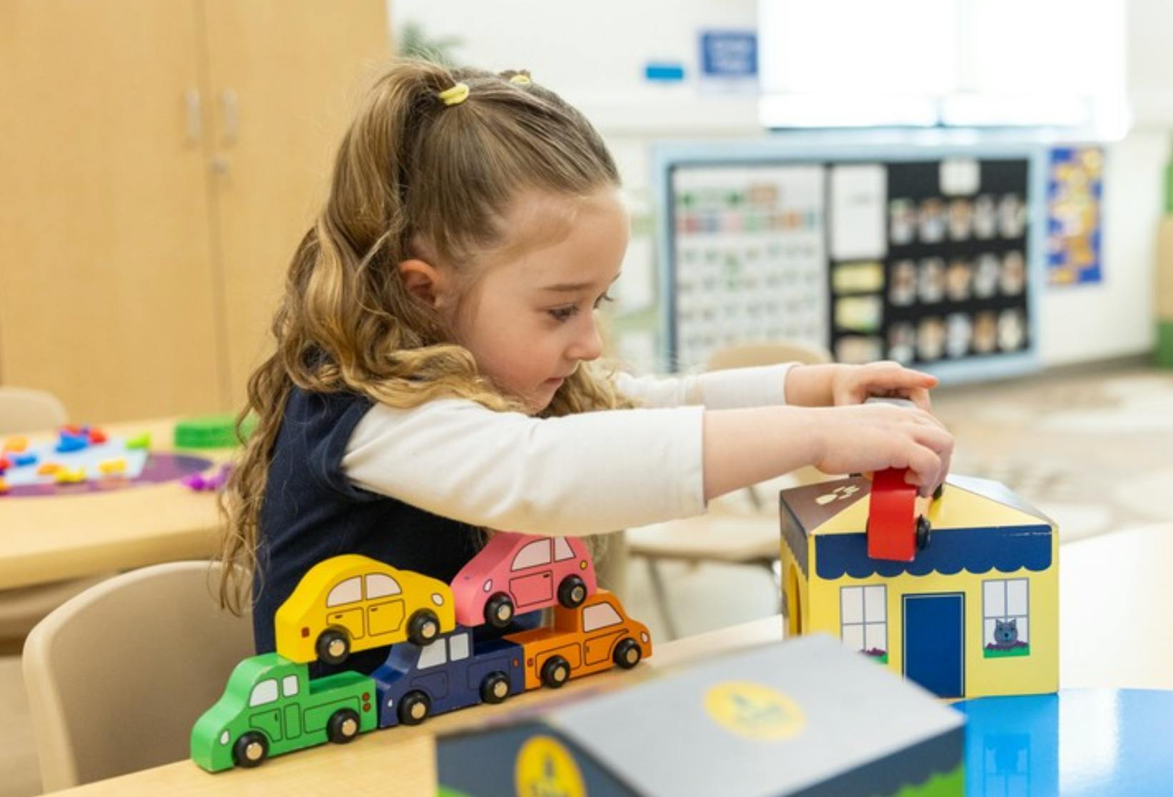 Child playing with a toy house and toy cars on a table in a classroom