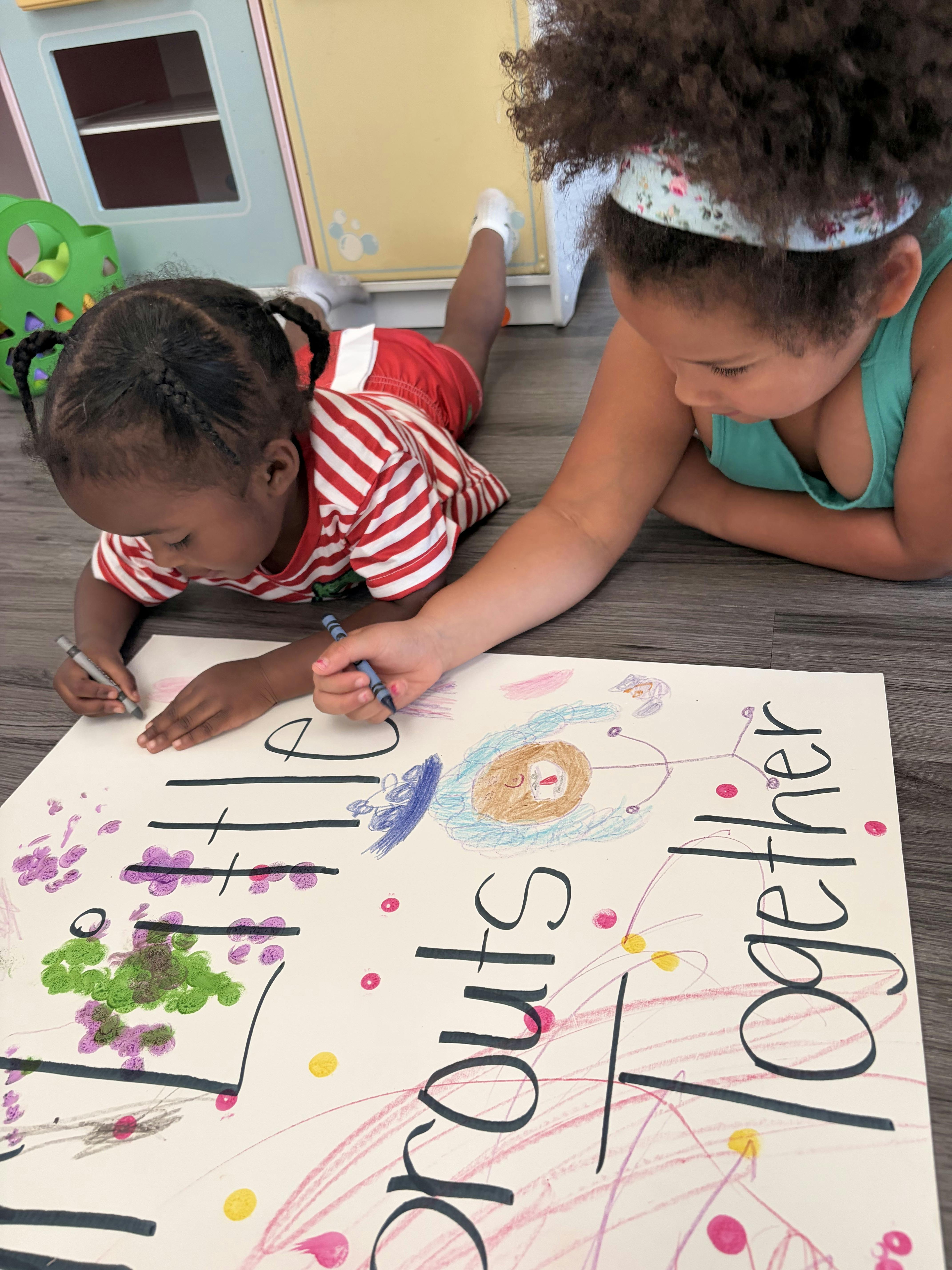 Two children lying on the floor drawing on a sign that says Little Sprouts Together