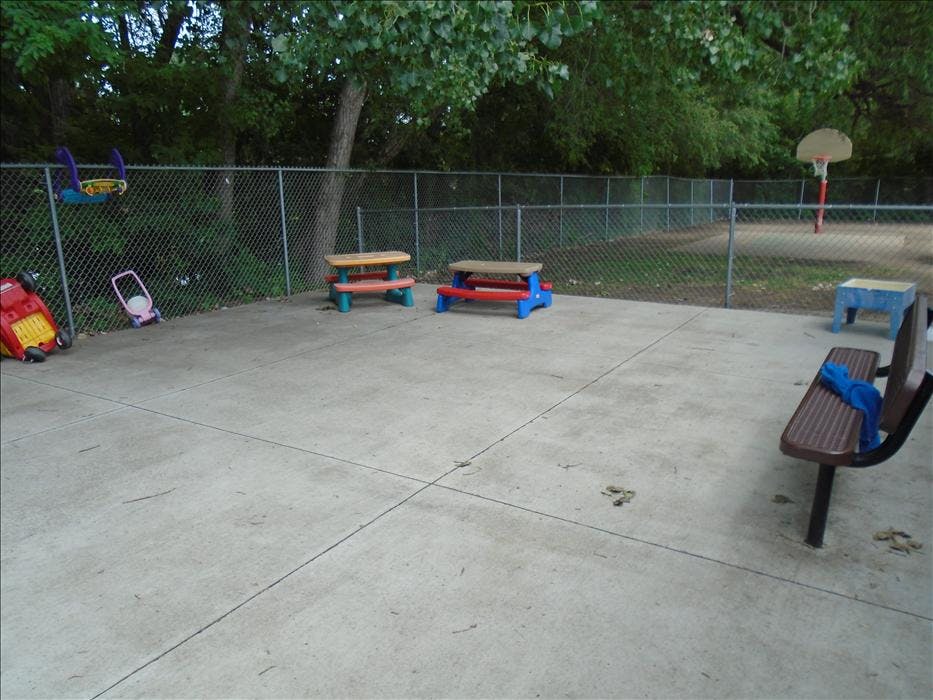 A fenced-in outdoor area with child-sized picnic tables and toys on a concrete slab