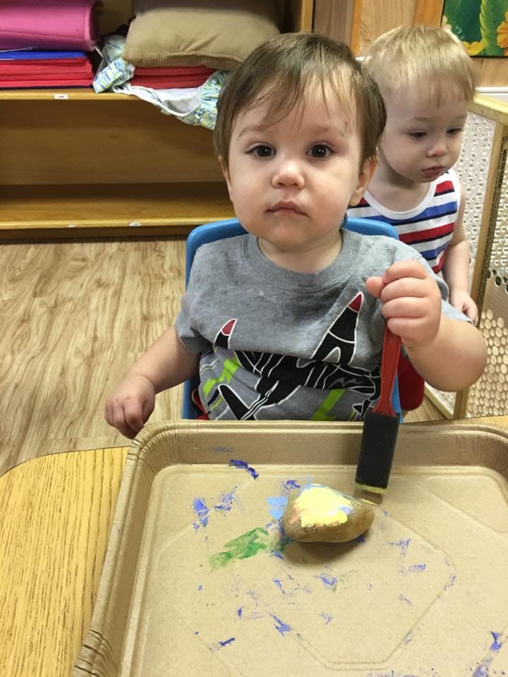 Child holding a paintbrush over a tray with paint and a rock