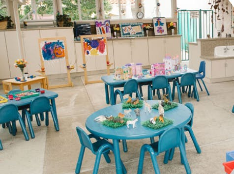 Classroom with small blue tables and chairs, easels with paintings