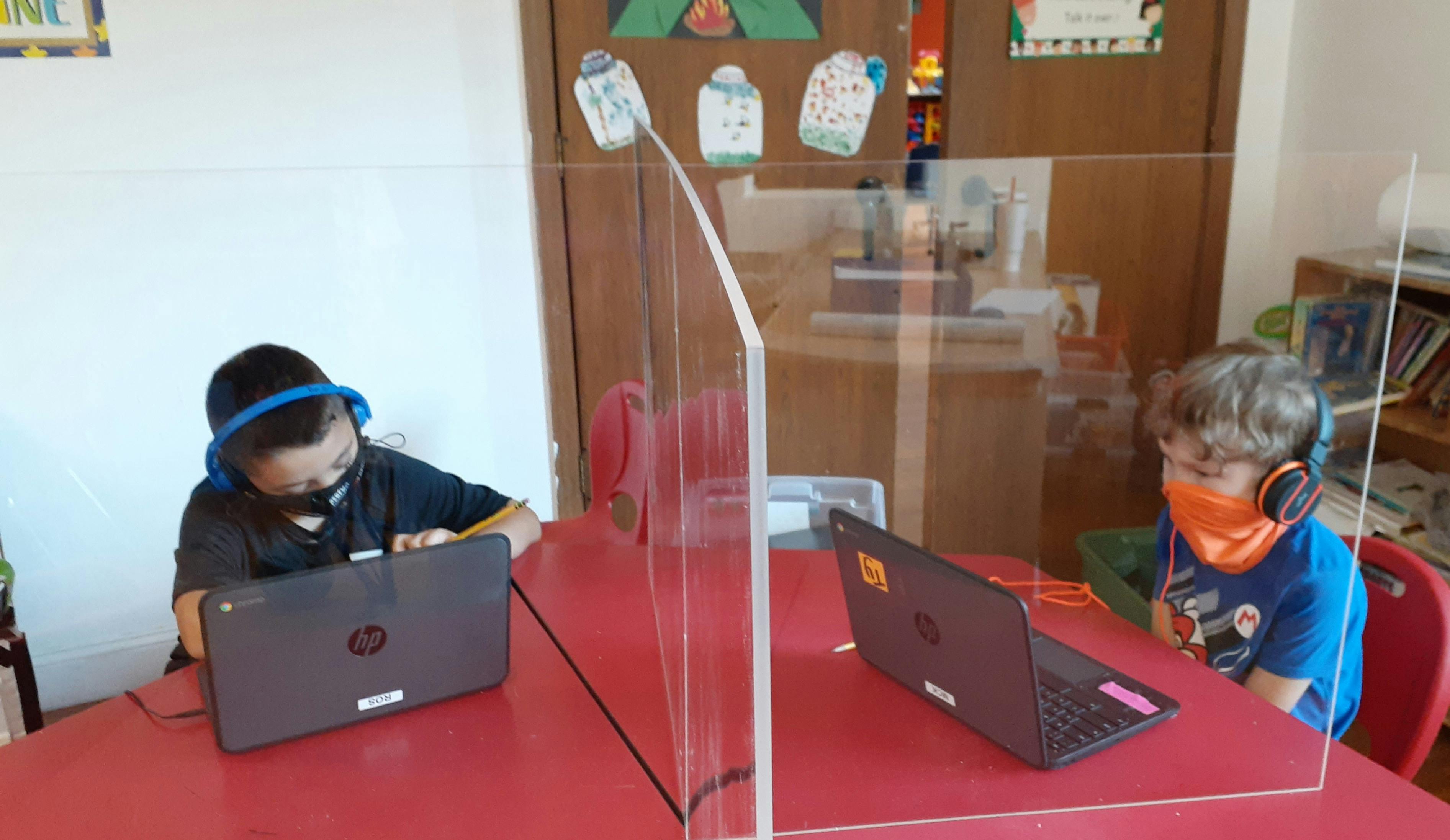 Two children with headphones and masks using laptops at a table with a clear divider