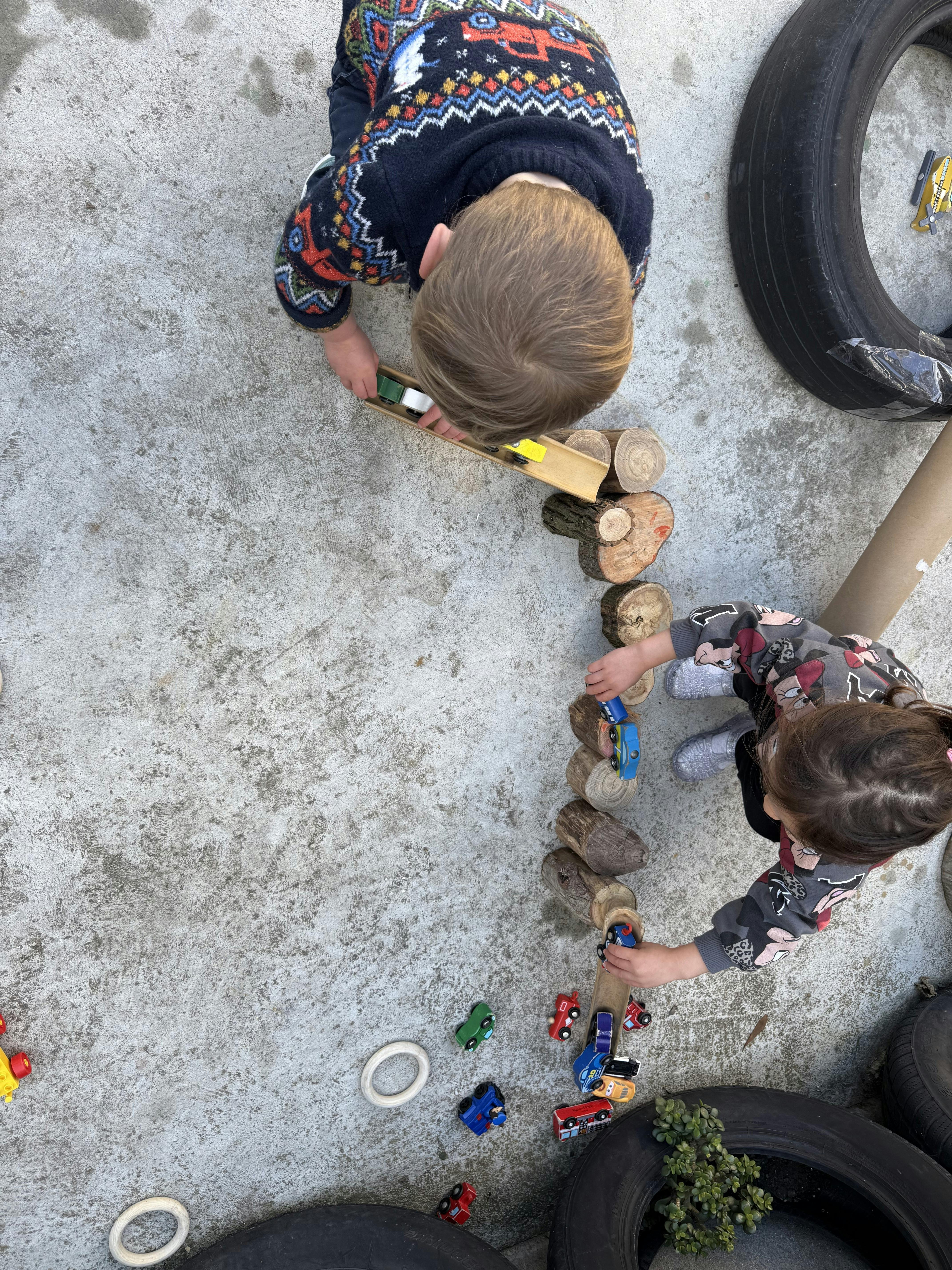 Two children playing with toy cars on a wooden ramp and logs