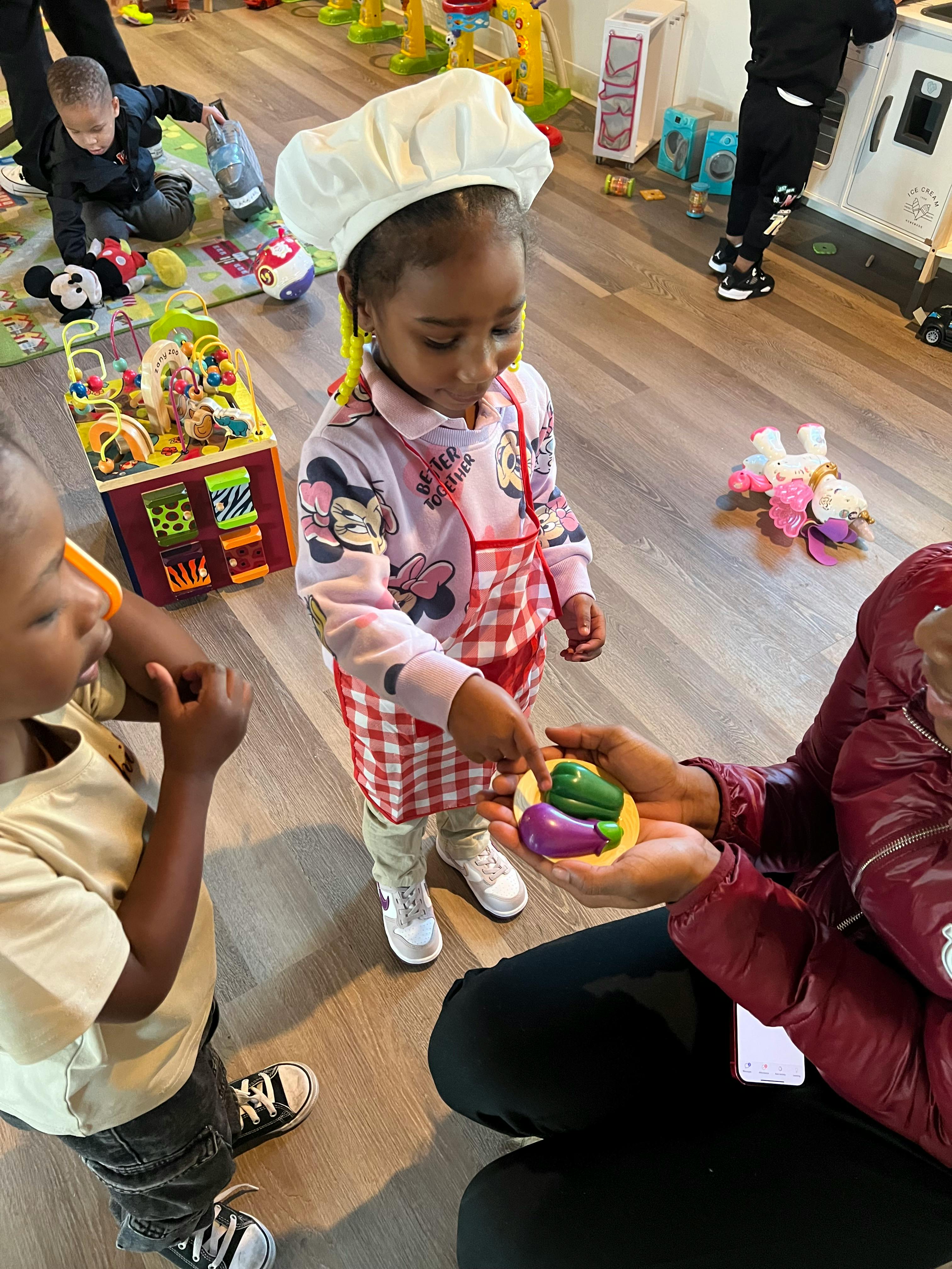 Child in chef hat pointing at toy vegetables held by an adult