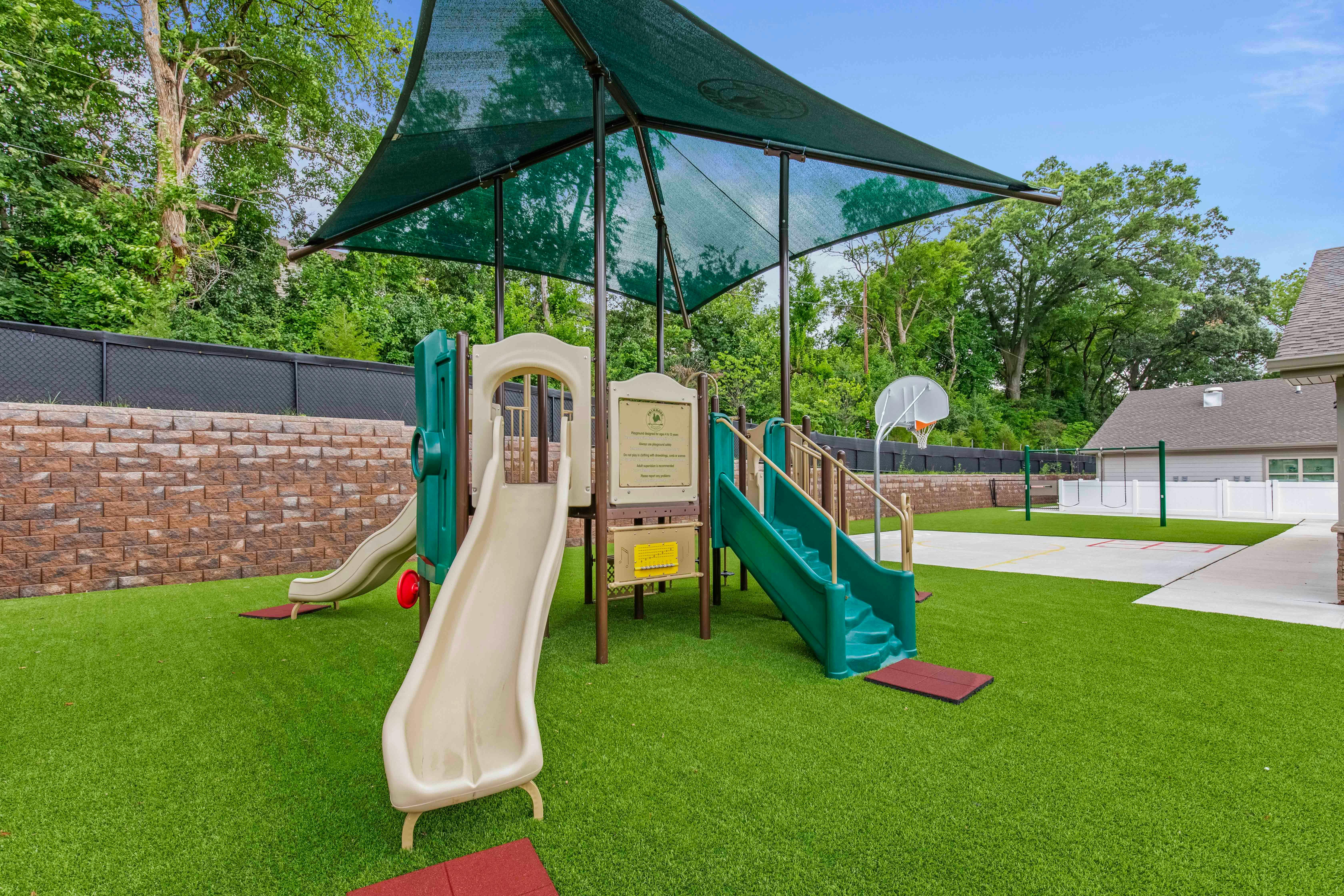 A playground with a green shade canopy over slides and stairs on artificial grass