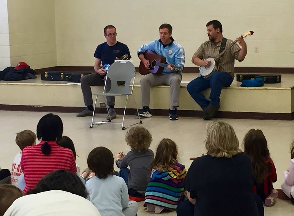 Three people playing musical instruments for an audience of children and adults