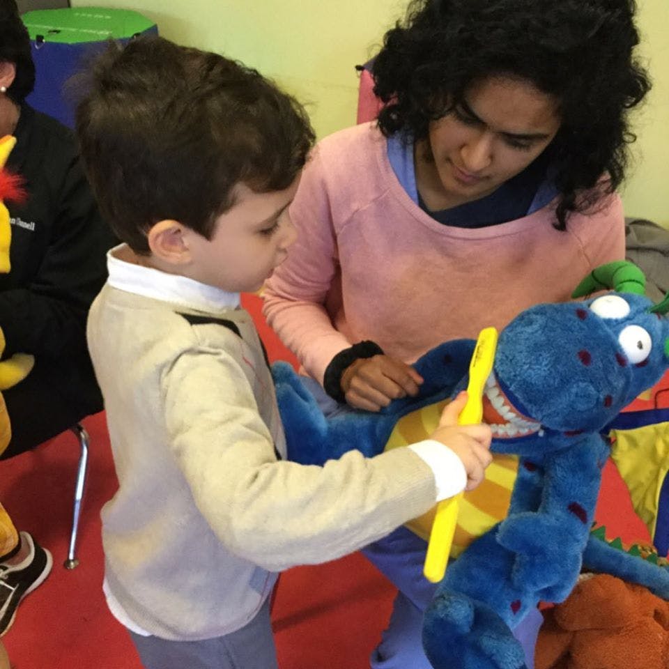 Child brushing teeth of a blue stuffed animal with a person watching