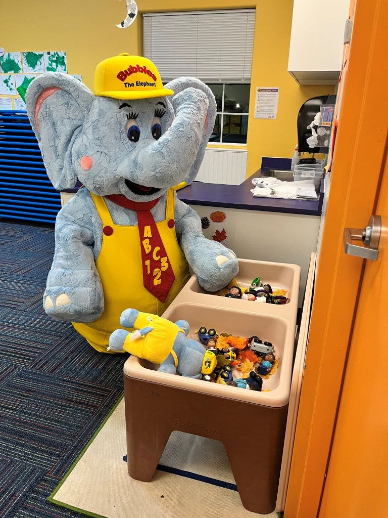 Elephant mascot in yellow overalls and hat next to a toy bin