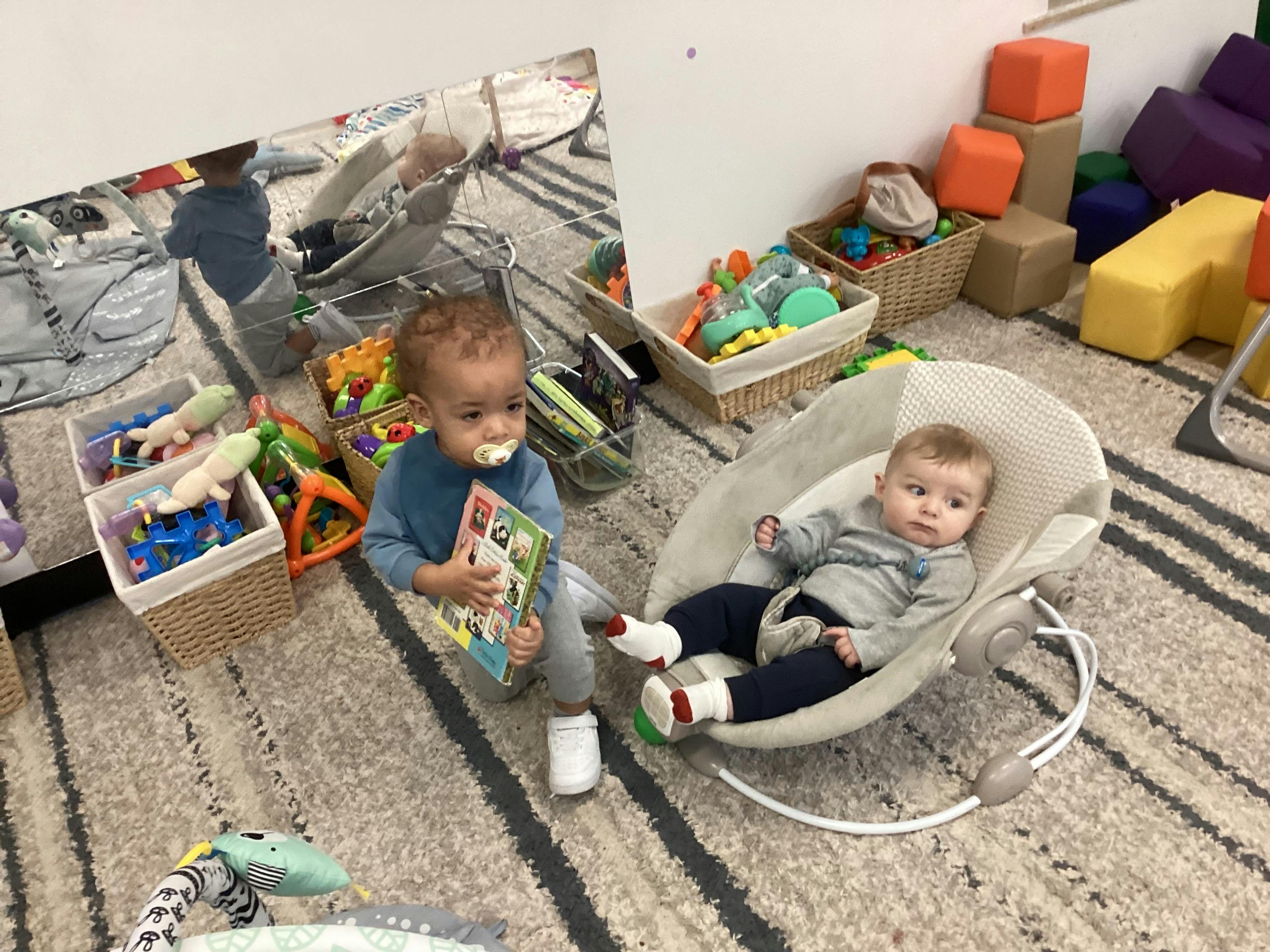 Child holding a book while sitting on a rug next to an infant in a bouncer