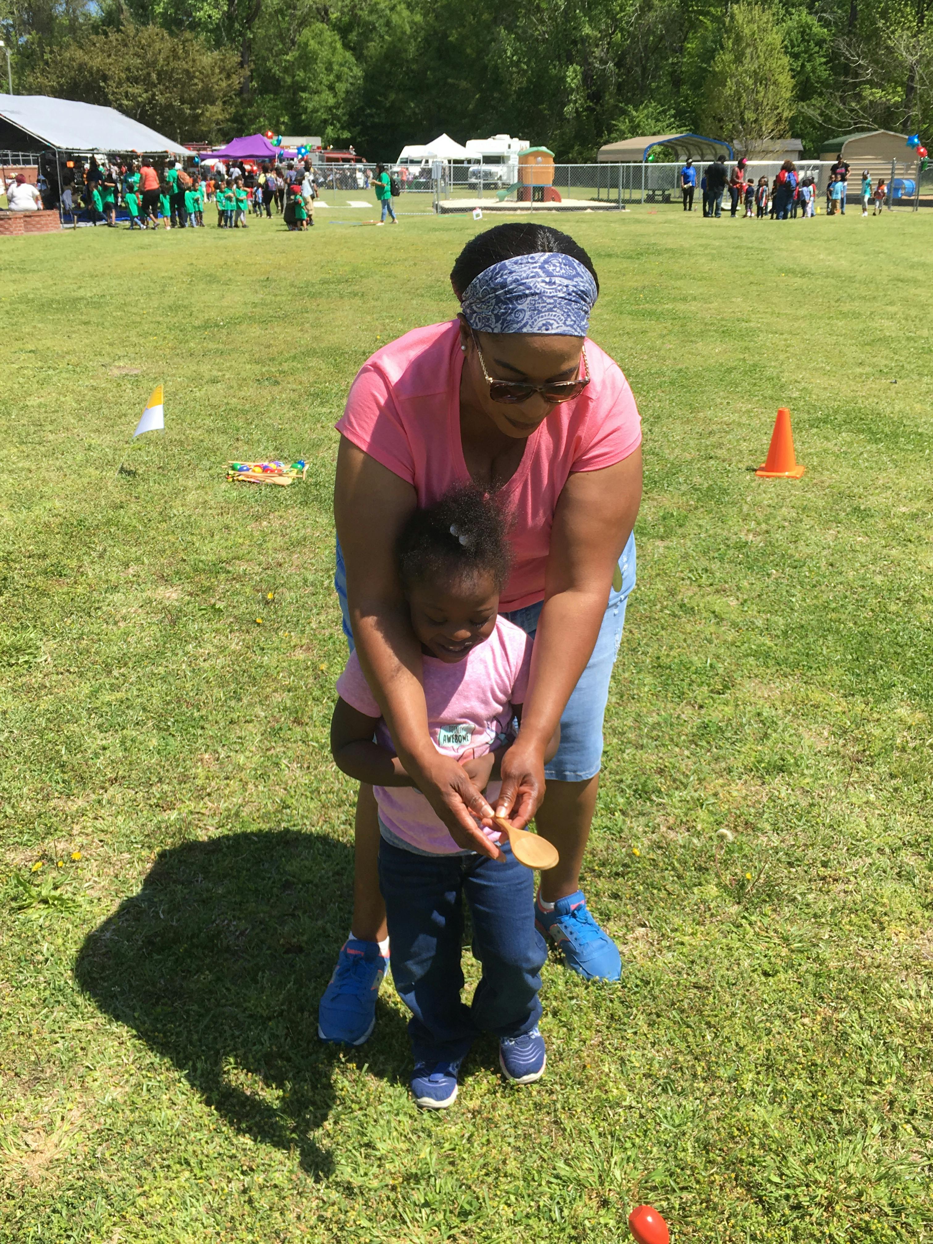 Person helping a child hold a wooden spoon on a grassy field