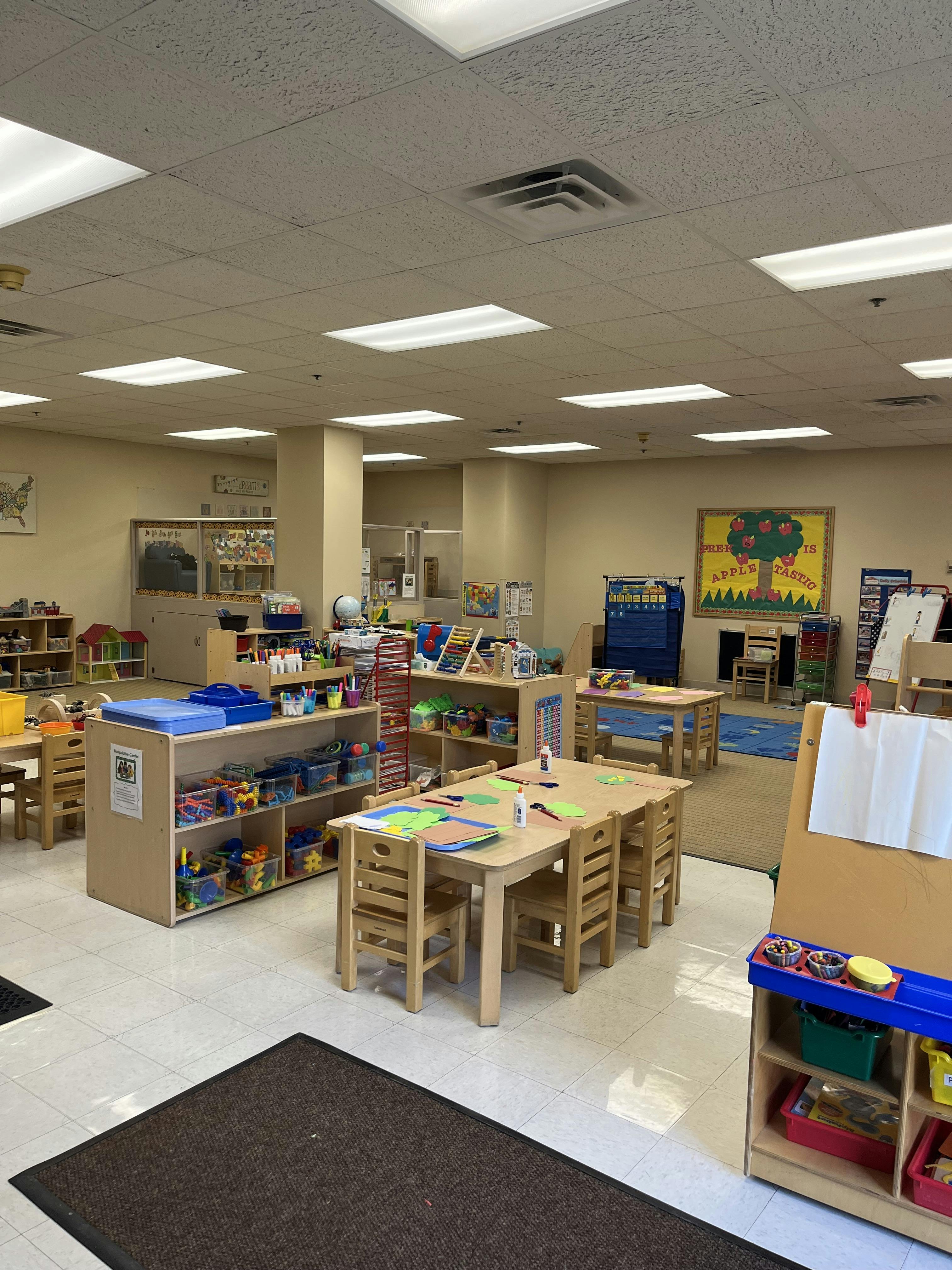 An empty classroom with small tables and chairs, shelves filled with toys