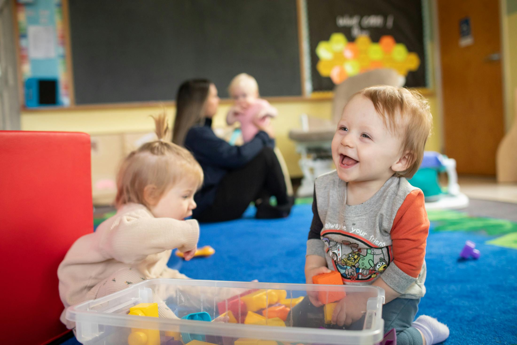 Two children playing with toys, person holding a child in background