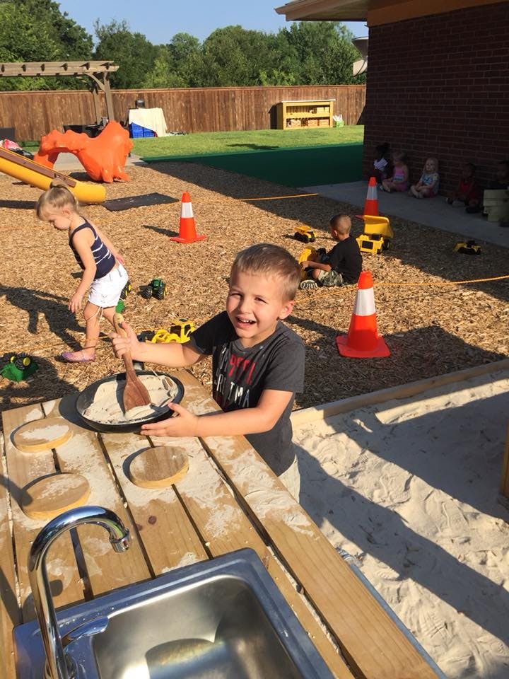 Child playing with sand and a wooden spoon at an outdoor sink