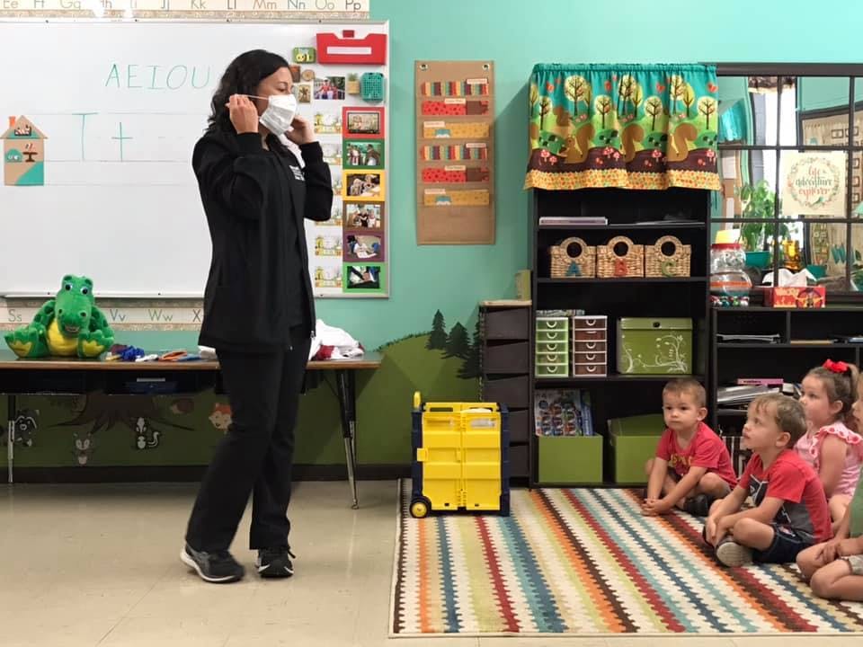 Person in a classroom putting on a face mask while children sit on a rug