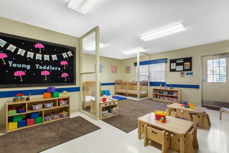 Toddler classroom with cribs, play tables, and a blackboard that says "Young Toddlers"