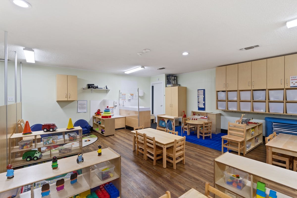 Empty preschool classroom with small tables, chairs, and shelving with toys