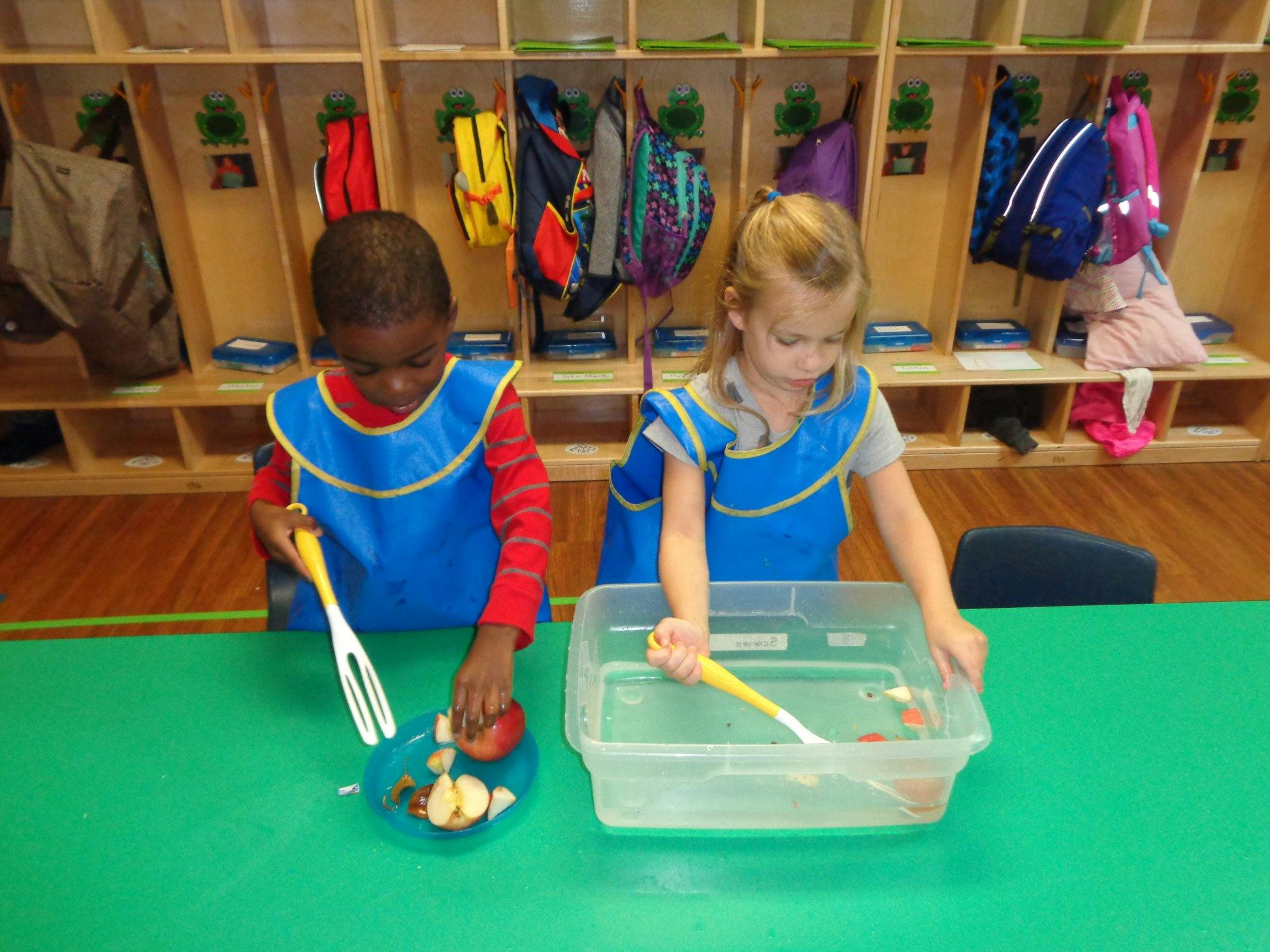 Two children in aprons playing with food and water in a water bin in a classroom