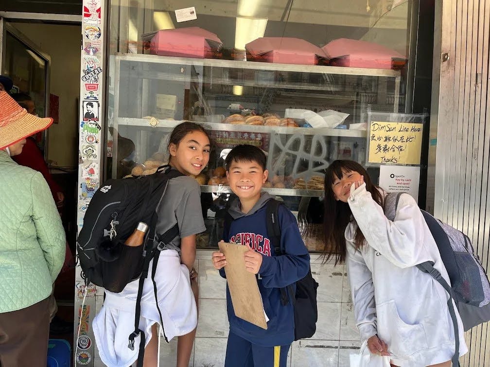 Three children with backpacks standing in front of a bakery window