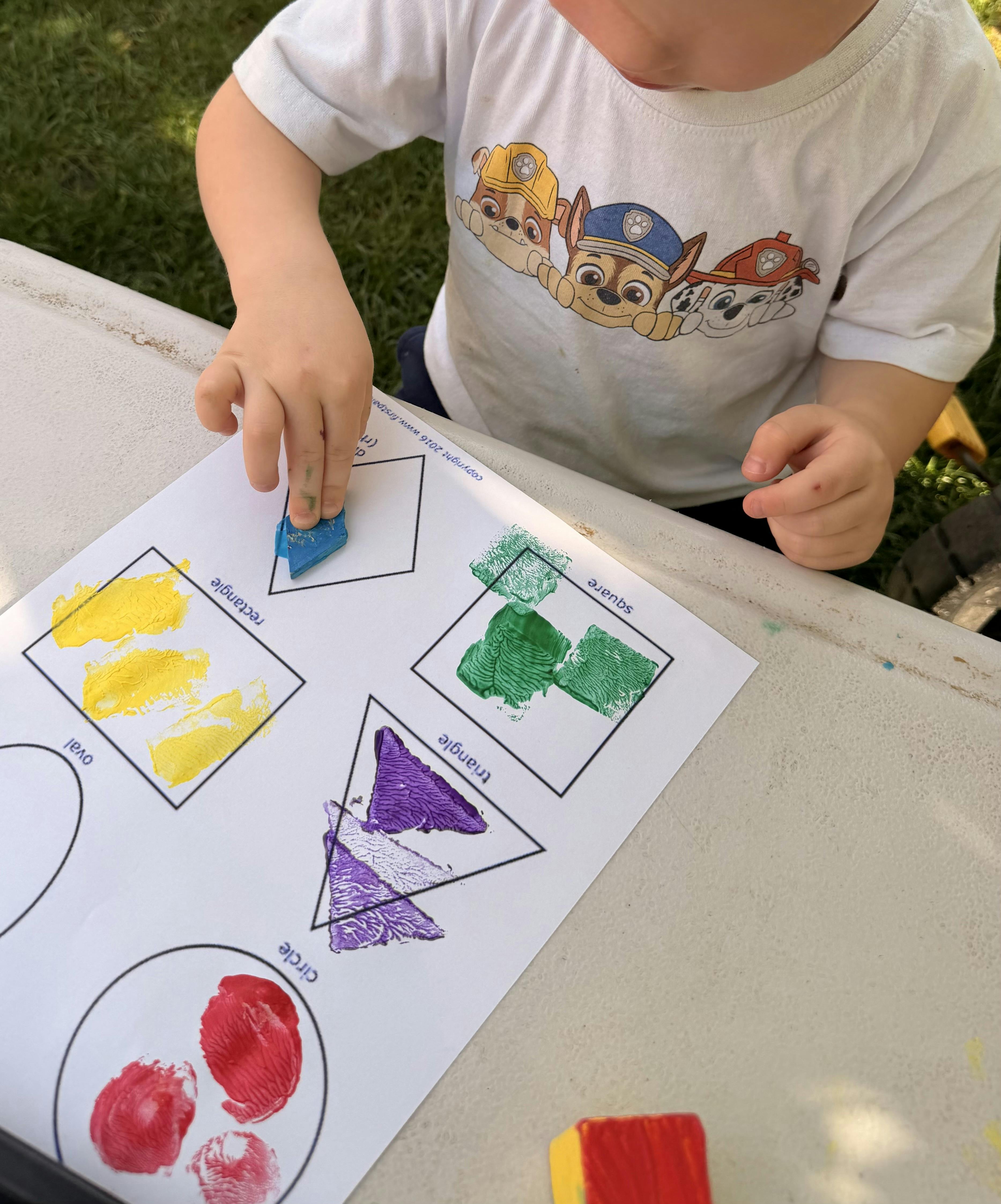 Child's hands pressing a blue block onto paper with painted shapes