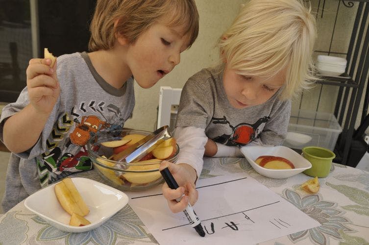 Two children at a table, one drawing on paper, the other holding food