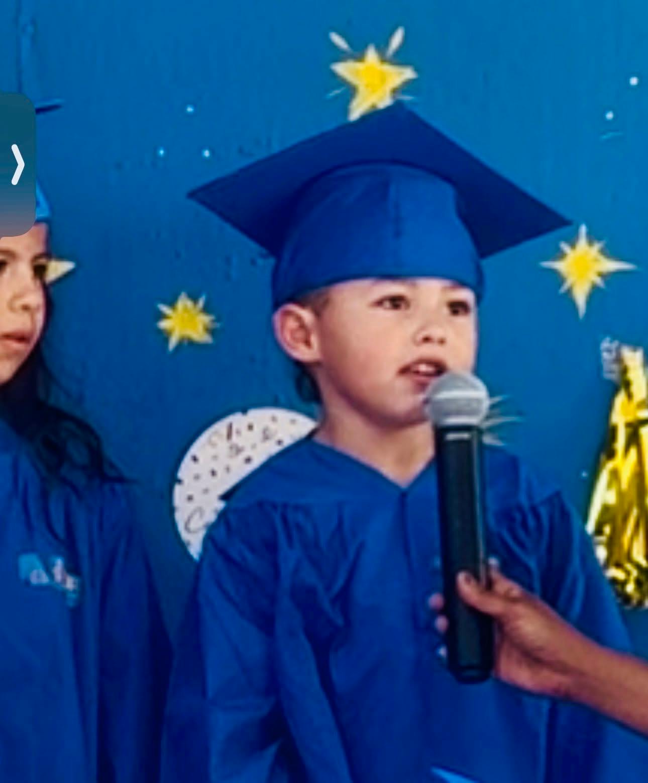 Child in blue cap and gown speaking into a microphone with another child nearby