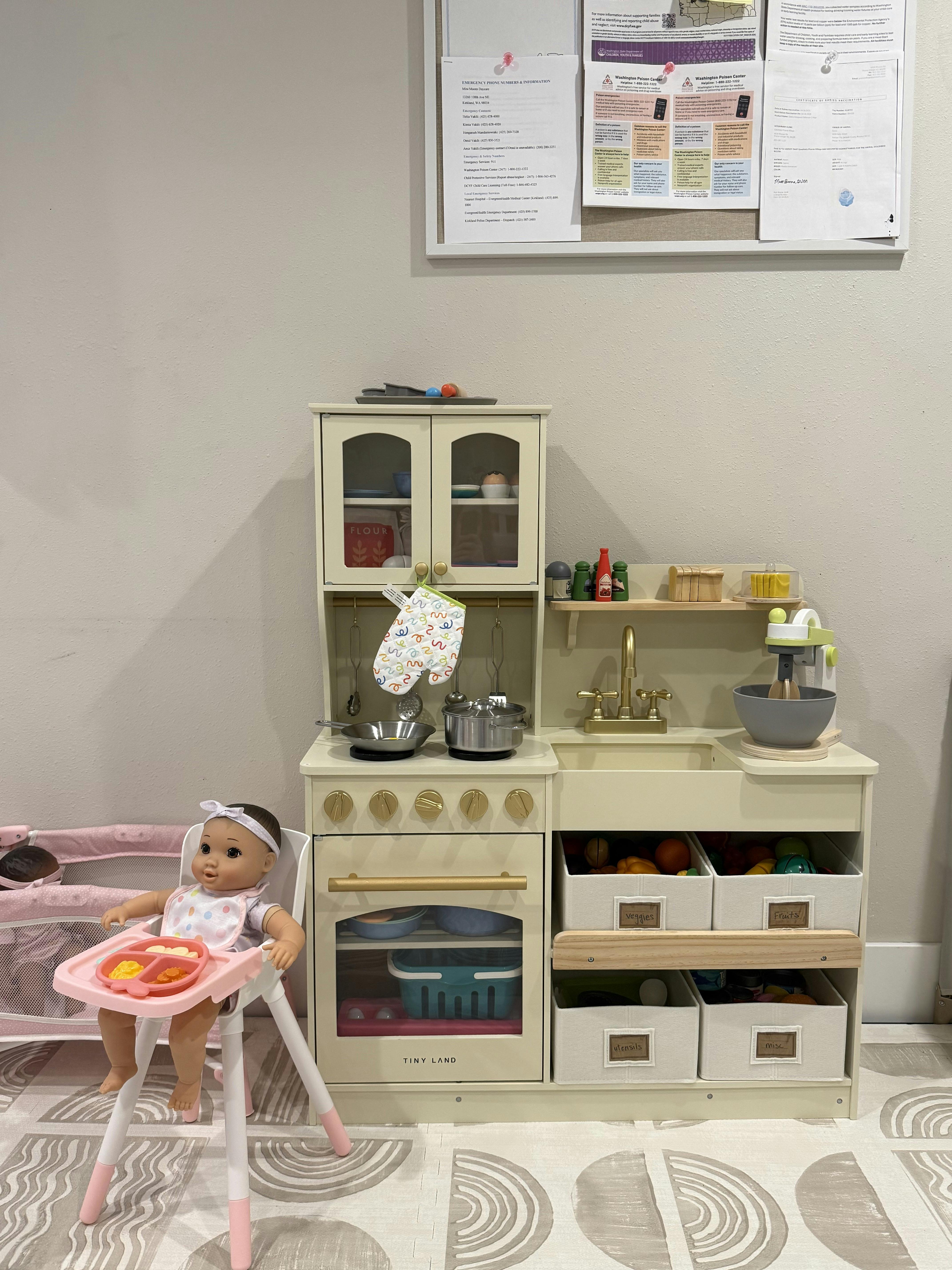 Doll in a highchair with a play food tray next to a play kitchen