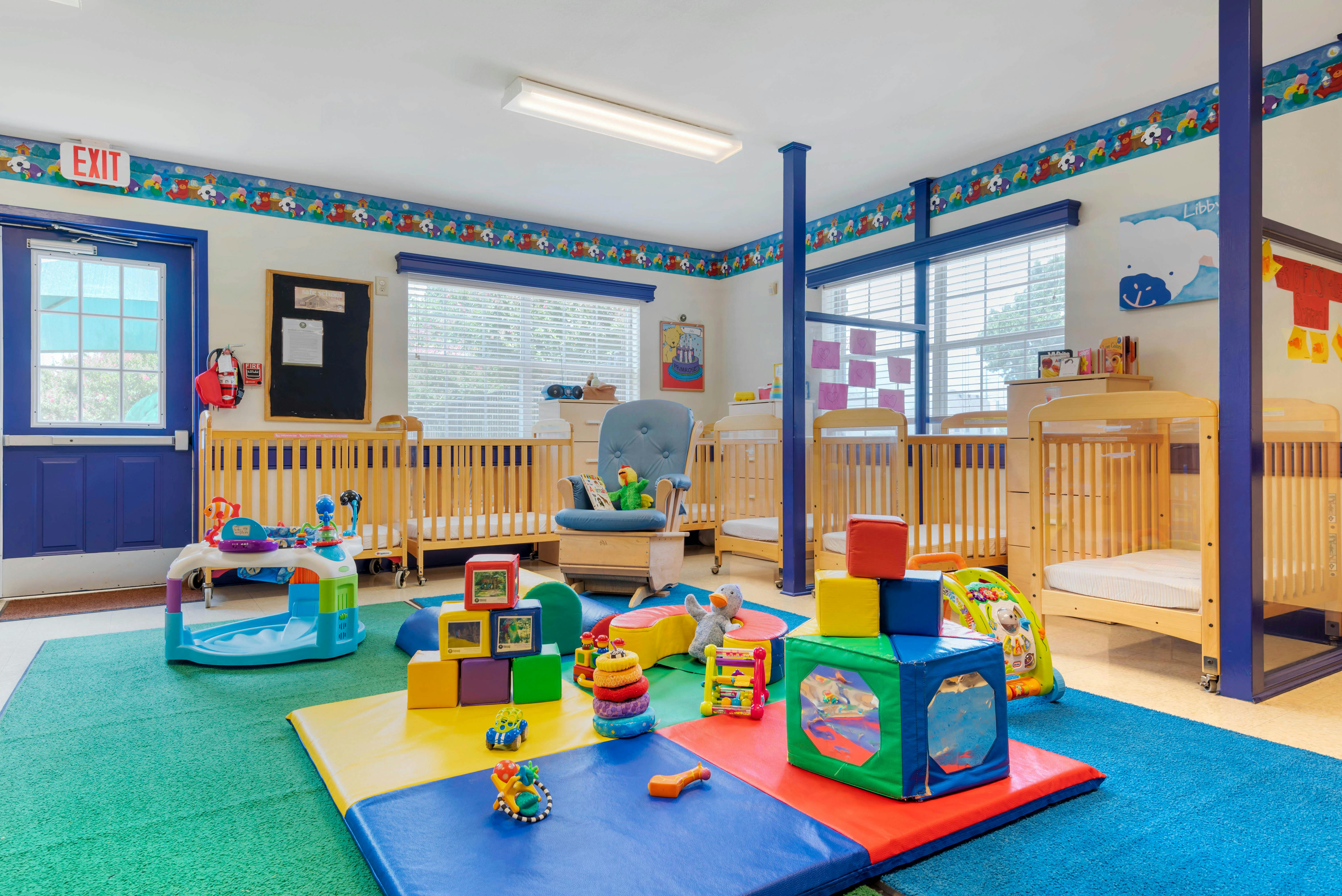 A colorful childcare room with cribs, playmats, toys, and a rocking chair
