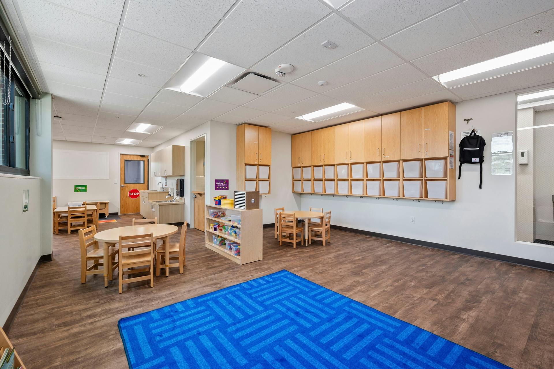 Empty classroom with small tables, chairs, cabinets, and a blue rug