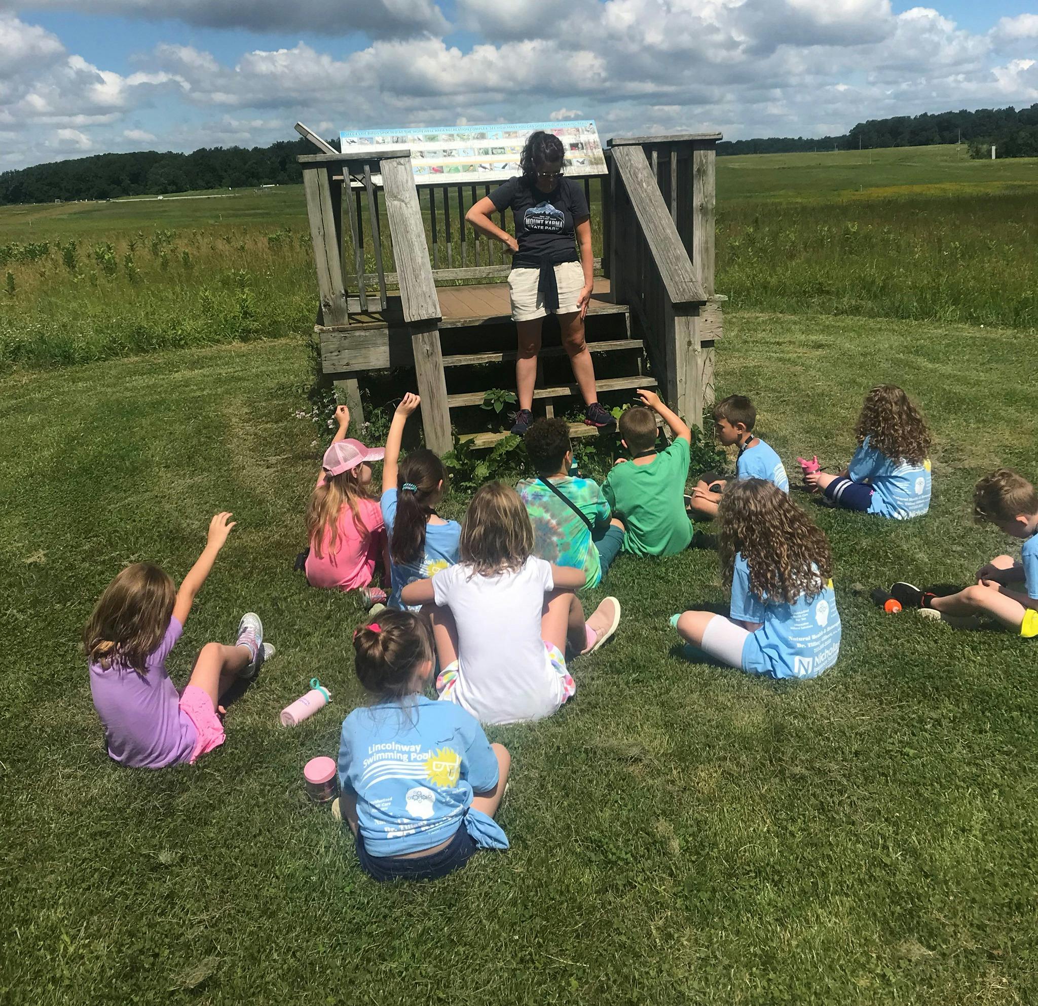 Person standing on wooden structure speaking to children seated on grass