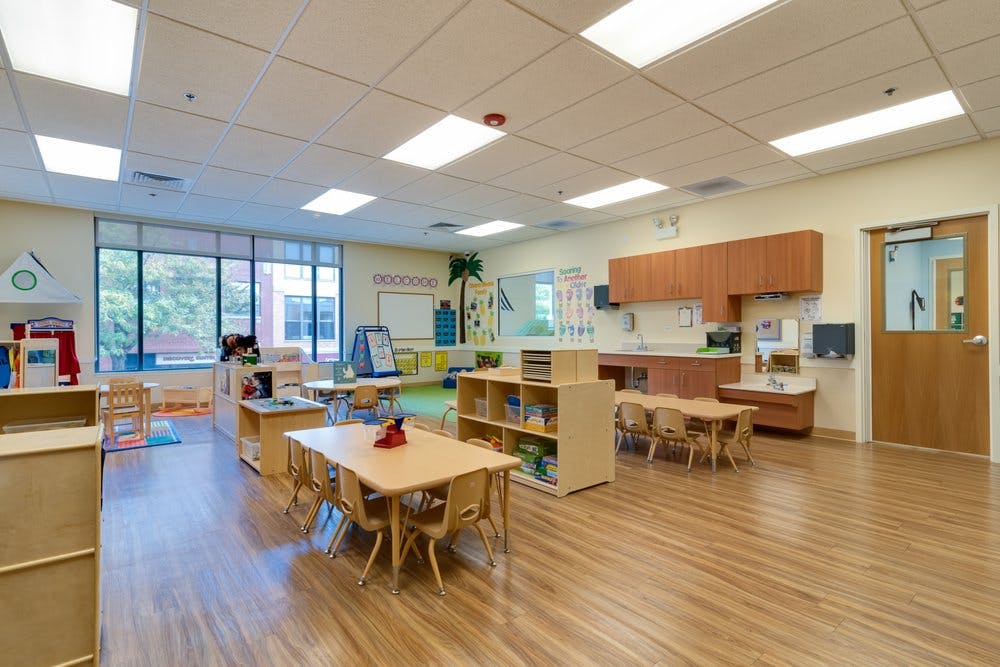 Empty classroom with small tables and chairs, storage shelves, and a counter area with large windows