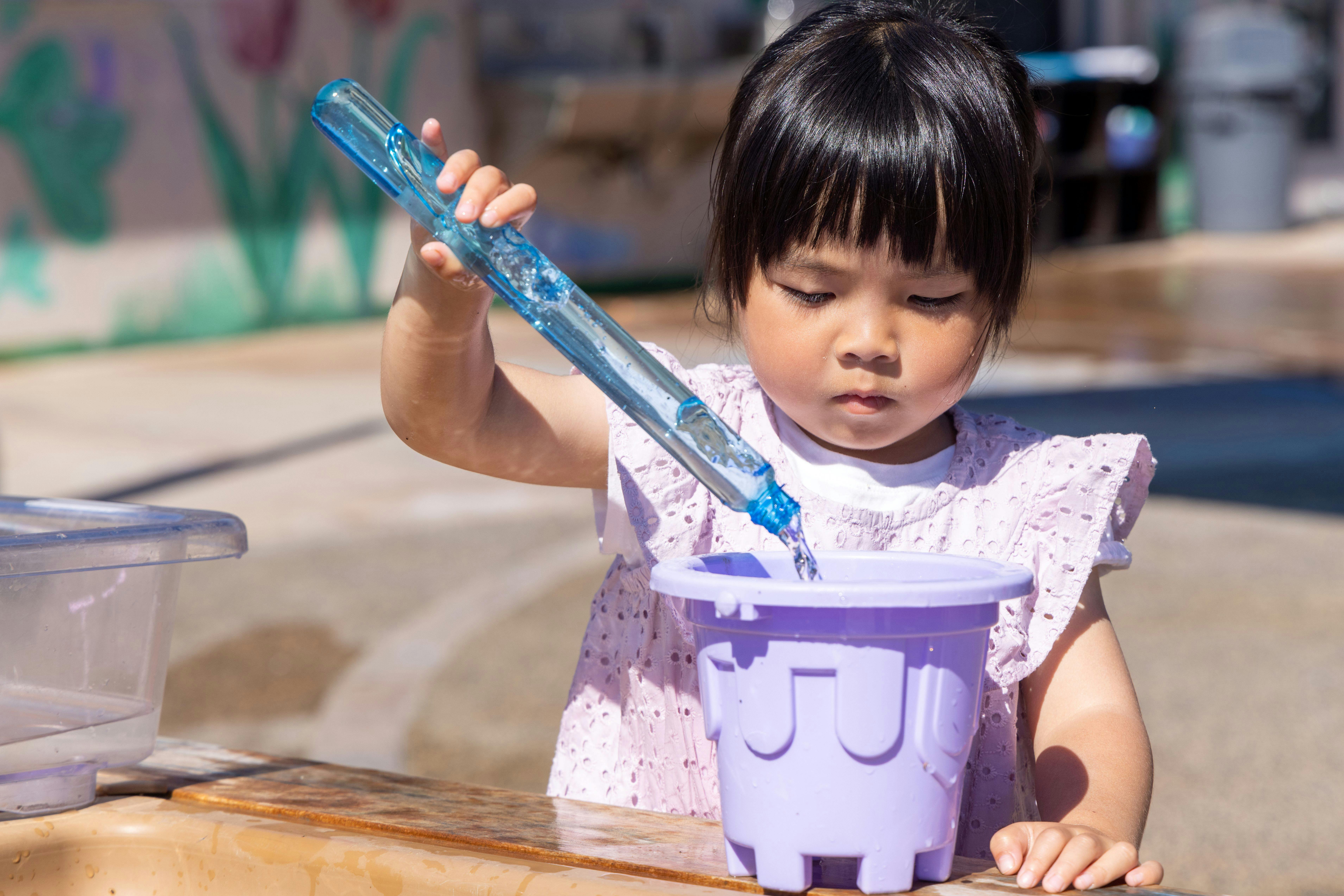 Child pouring water from a blue toy into a purple bucket