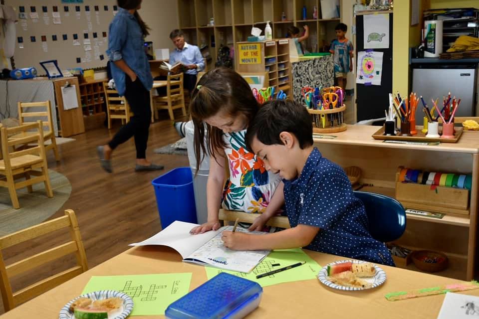Two children writing in a book at a table in a classroom