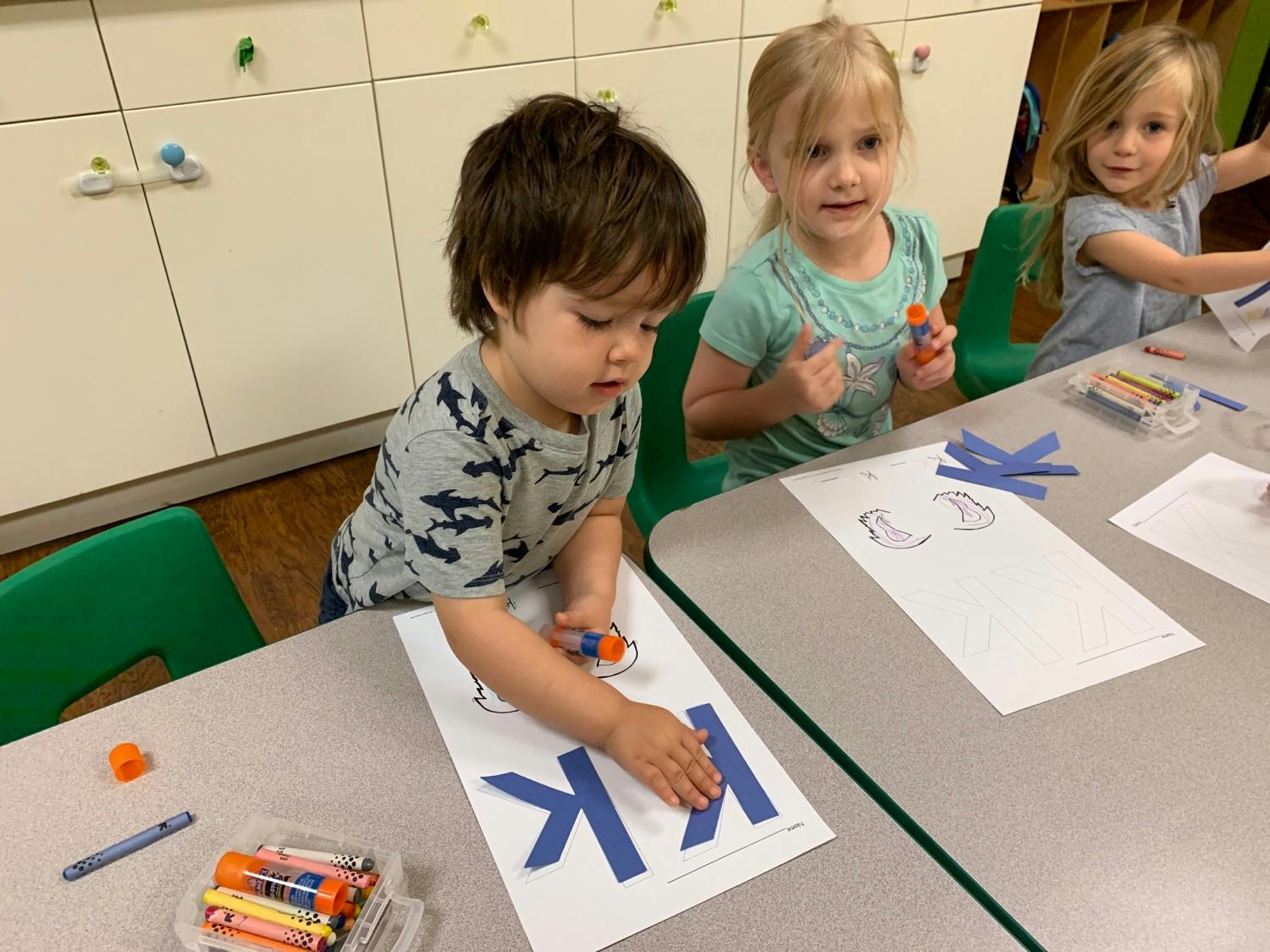 Three children at a table doing crafts, one child gluing blue letter K cutouts