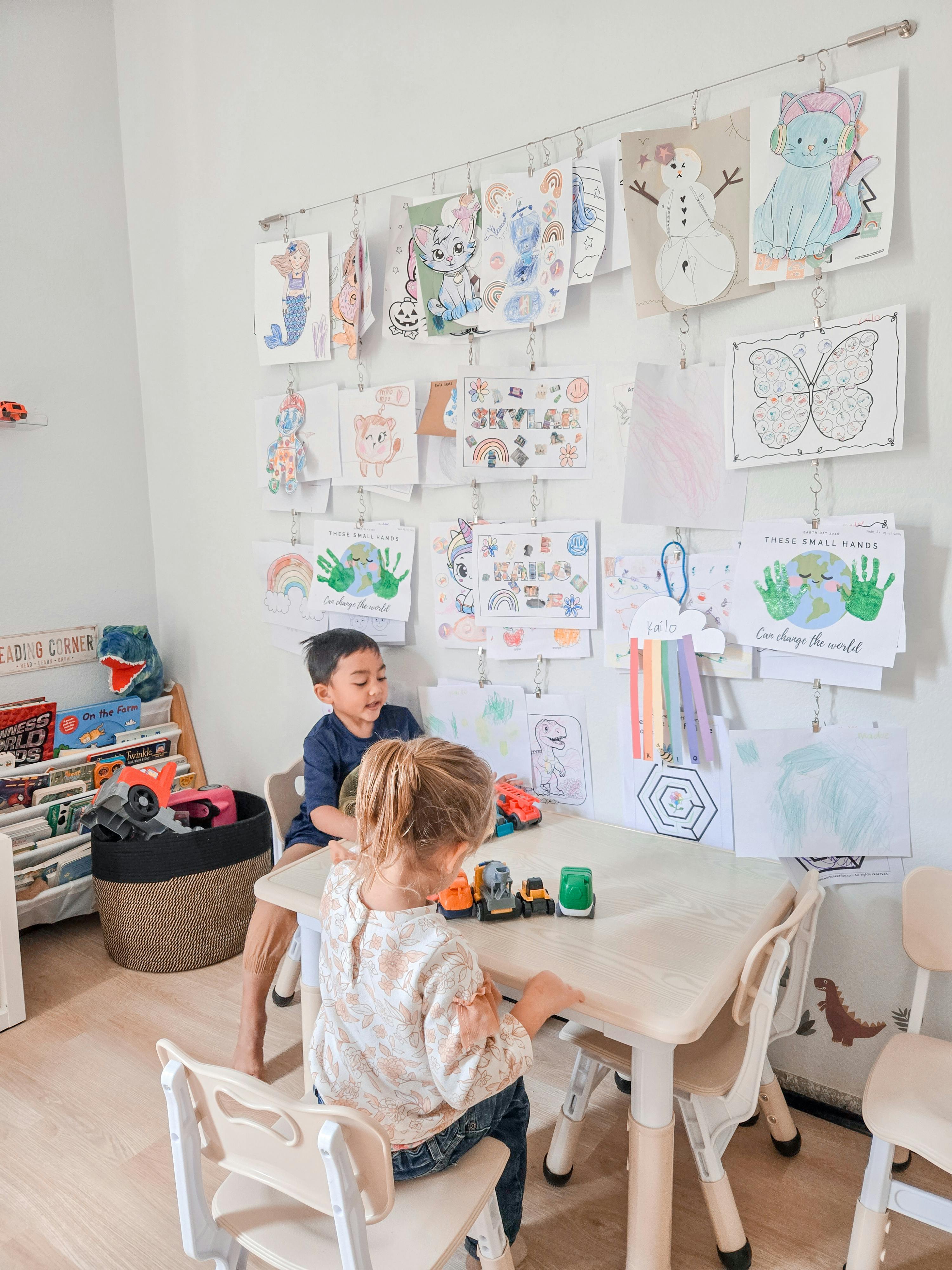 Two children playing with toy vehicles at a table in a room decorated with art