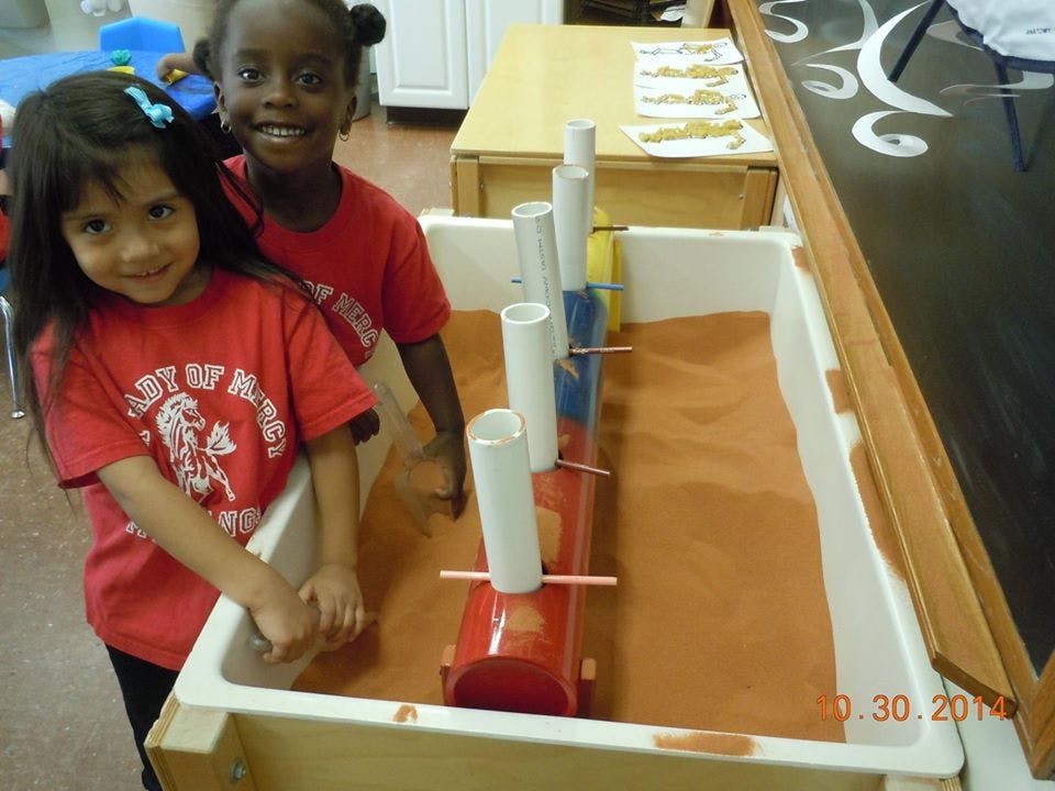 Two children standing next to a sand table with tubes and scoops