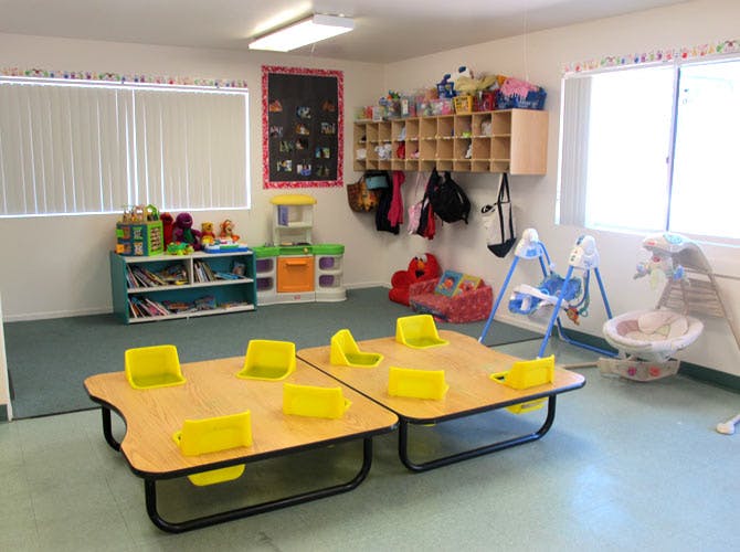 Classroom with small tables and chairs, shelves with toys, and baby swings