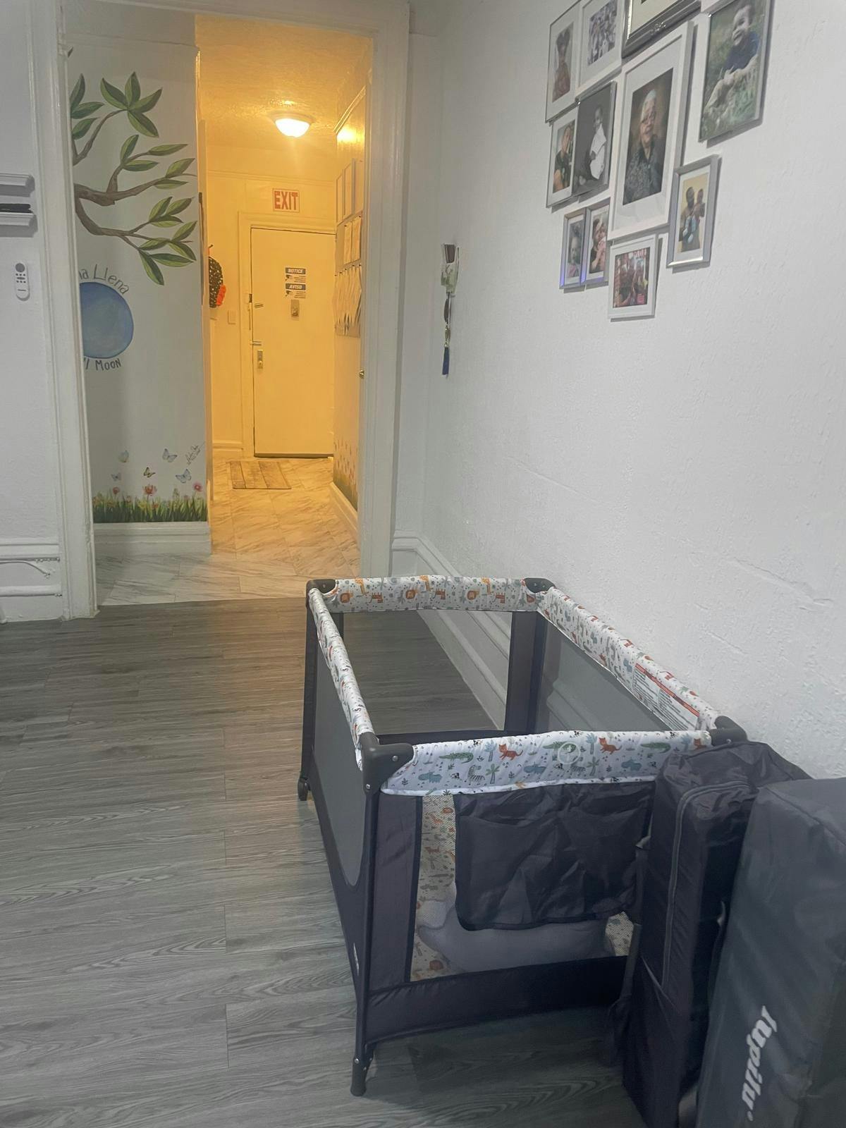 An empty portable playpen on a grey wood floor, with a doorway and a wall with family photos