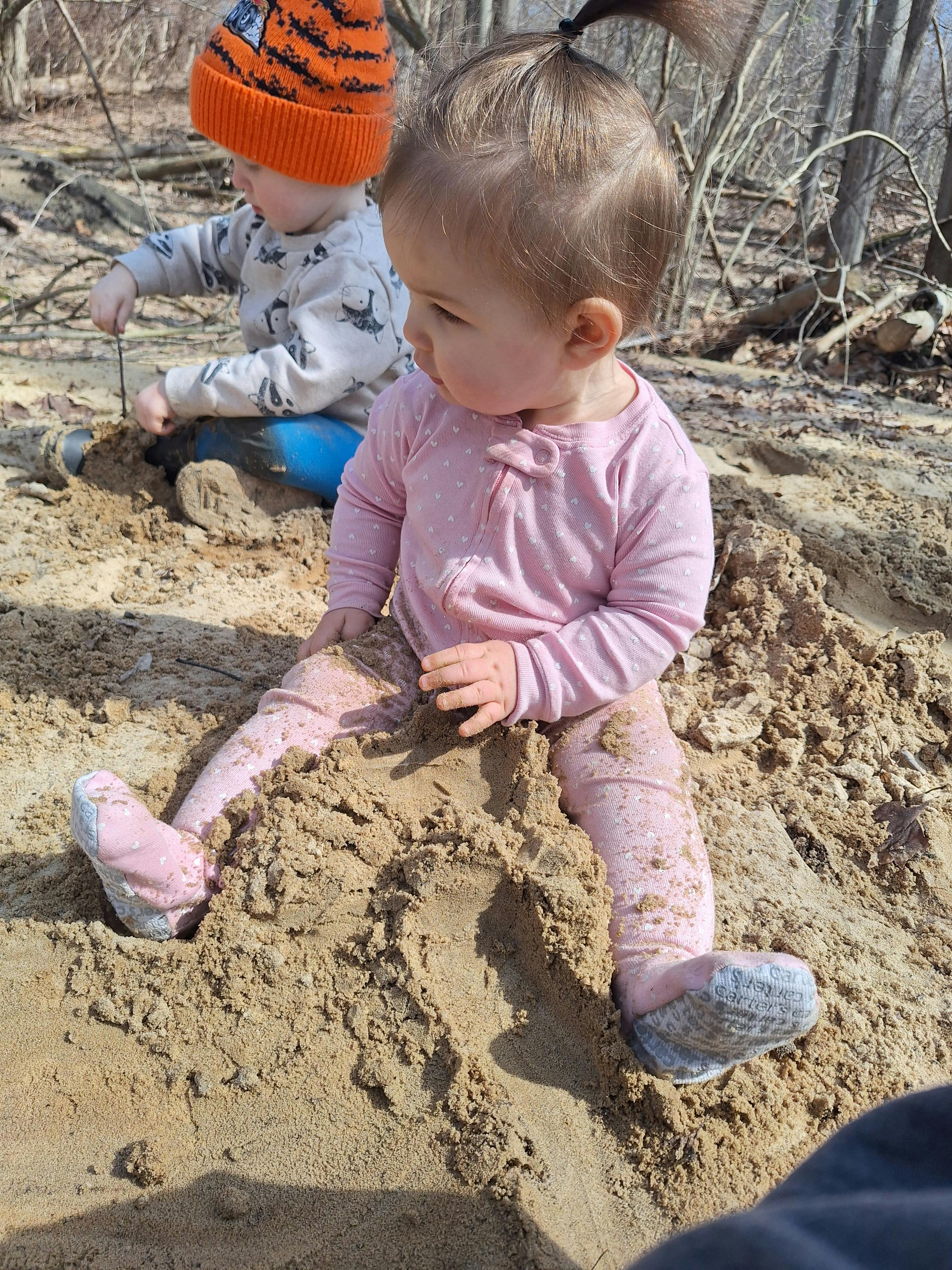Two children playing in a sandy area, one wearing pink clothing and the other an orange hat