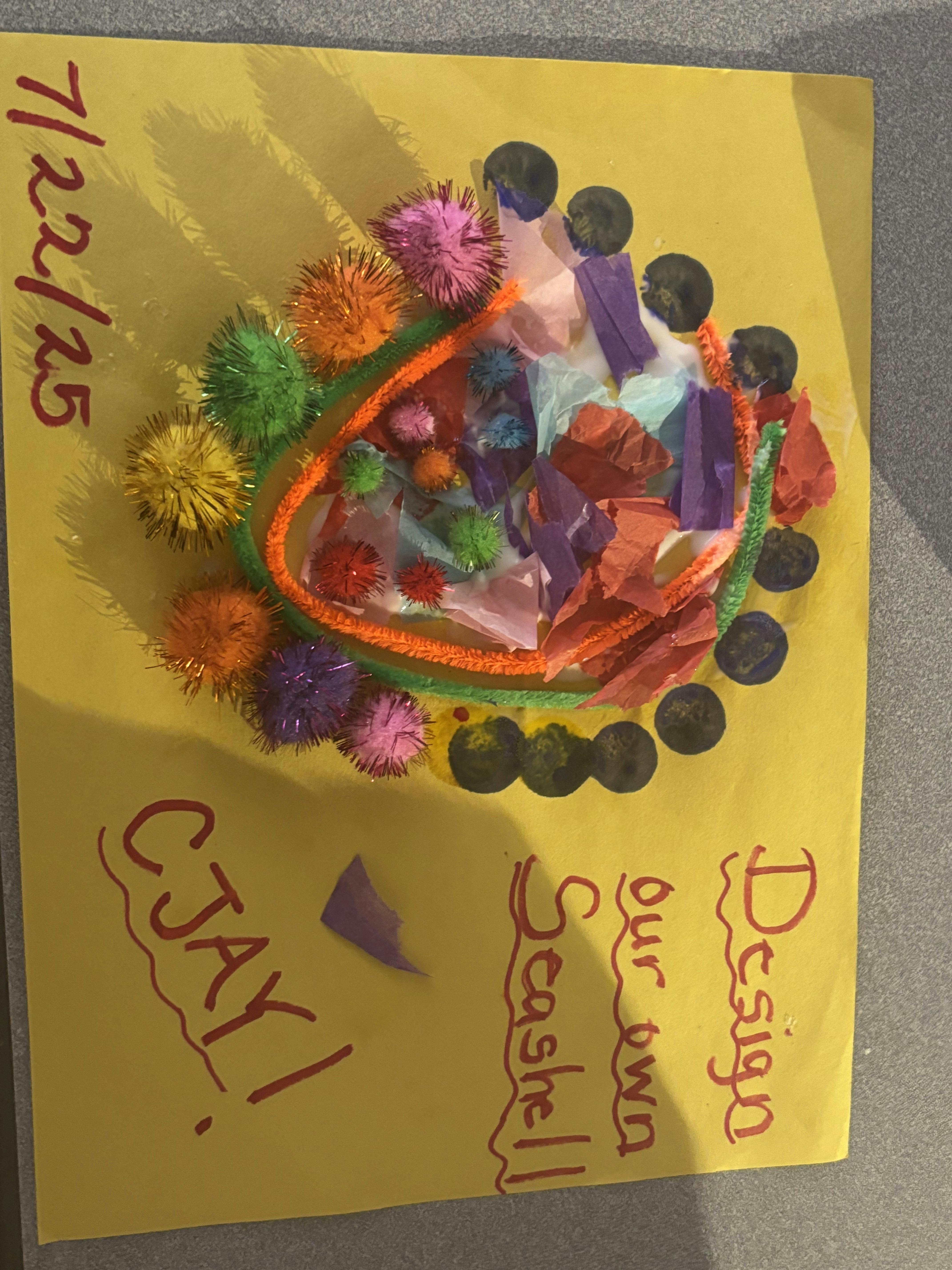 Child's craft project of a seashell shape decorated with pom-poms and tissue paper