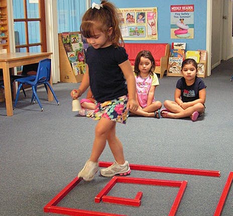 Child walking on red shapes on the floor, two children sitting in background