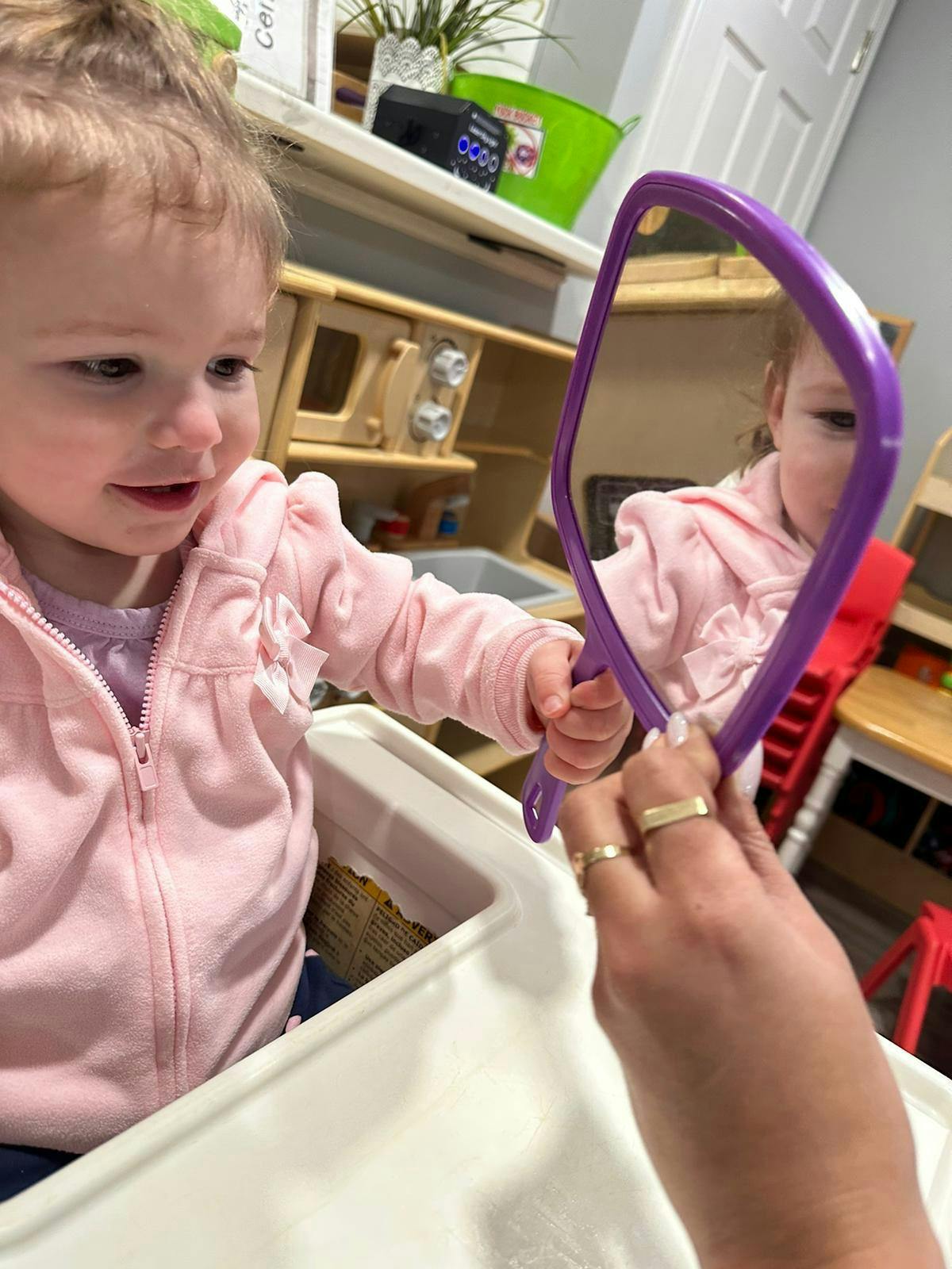 Child in high chair looking at reflection in a purple mirror held by an adult