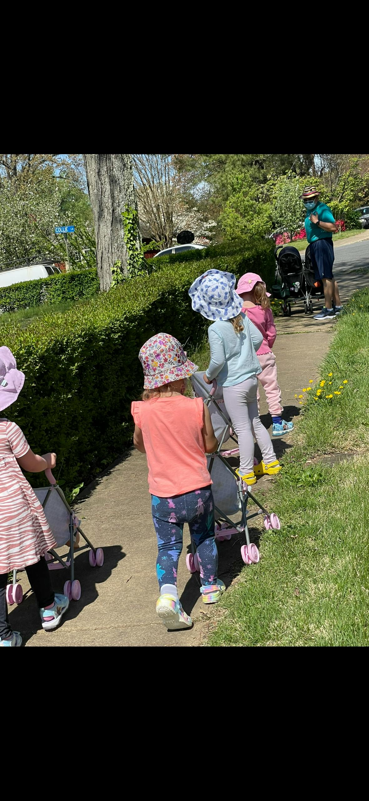 Children pushing toy strollers on a sidewalk with an adult in the background