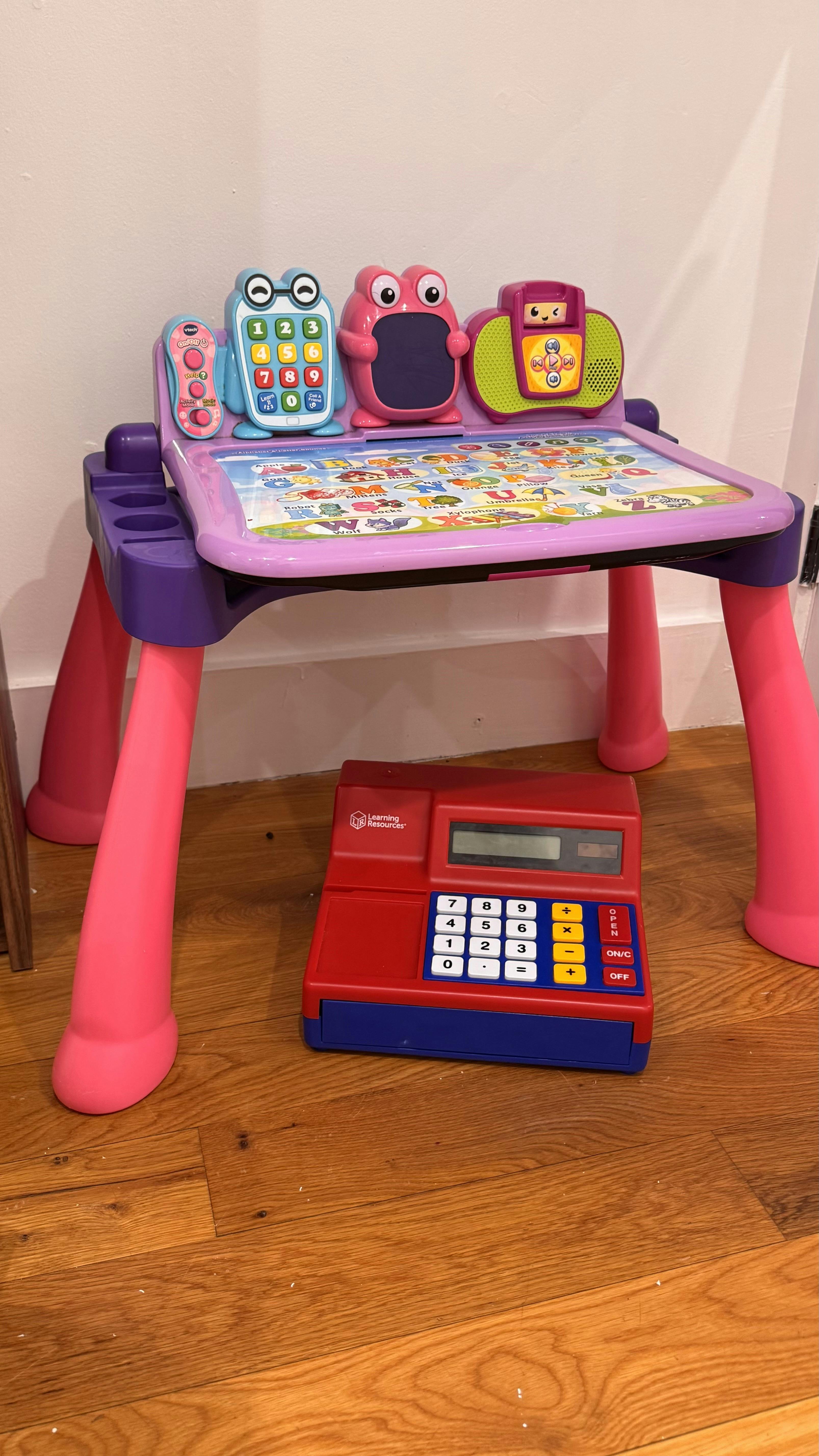 A purple and pink toy learning desk with a red and blue toy cash register underneath