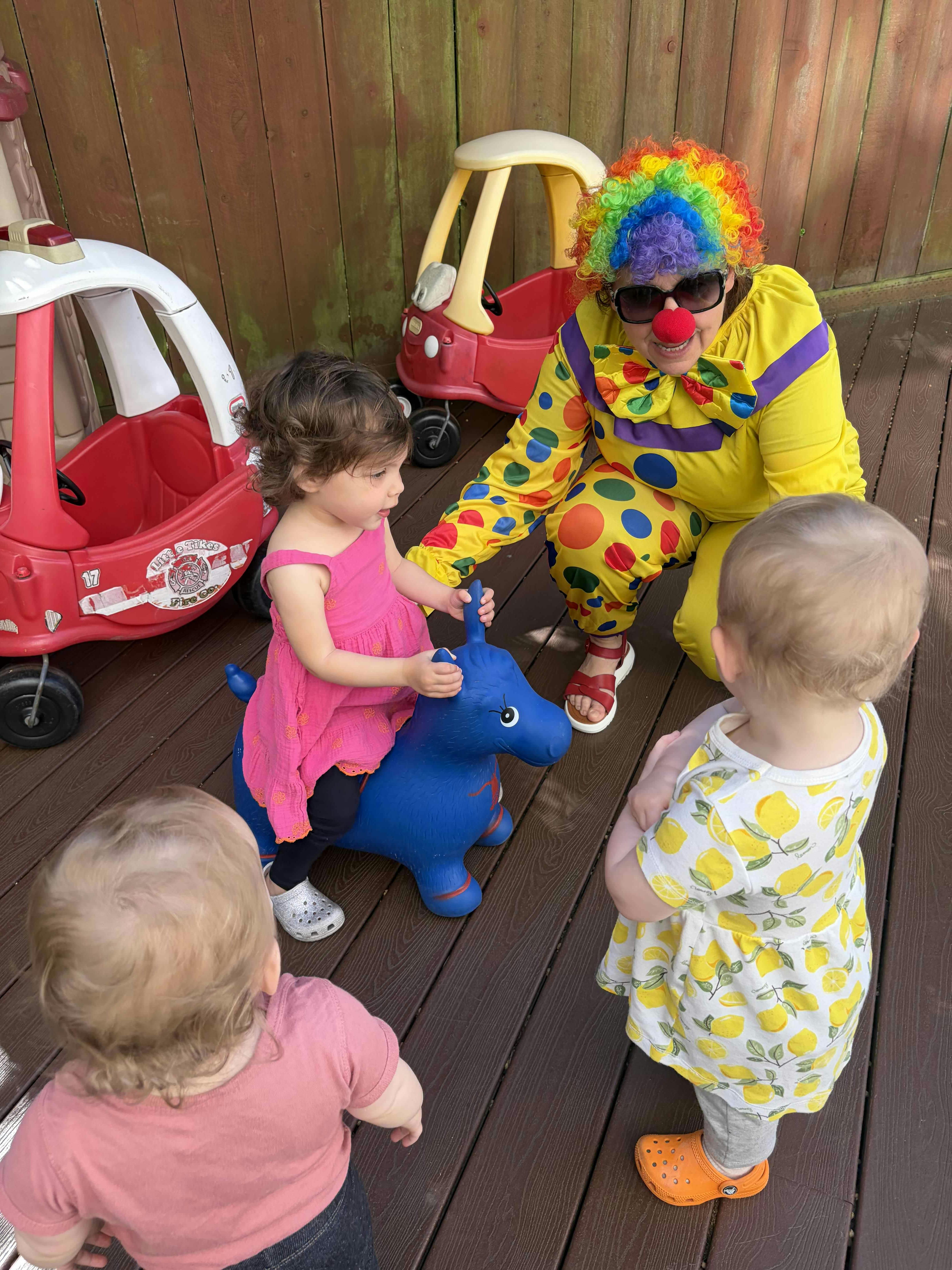 Person in a clown costume interacting with three children on a wooden deck