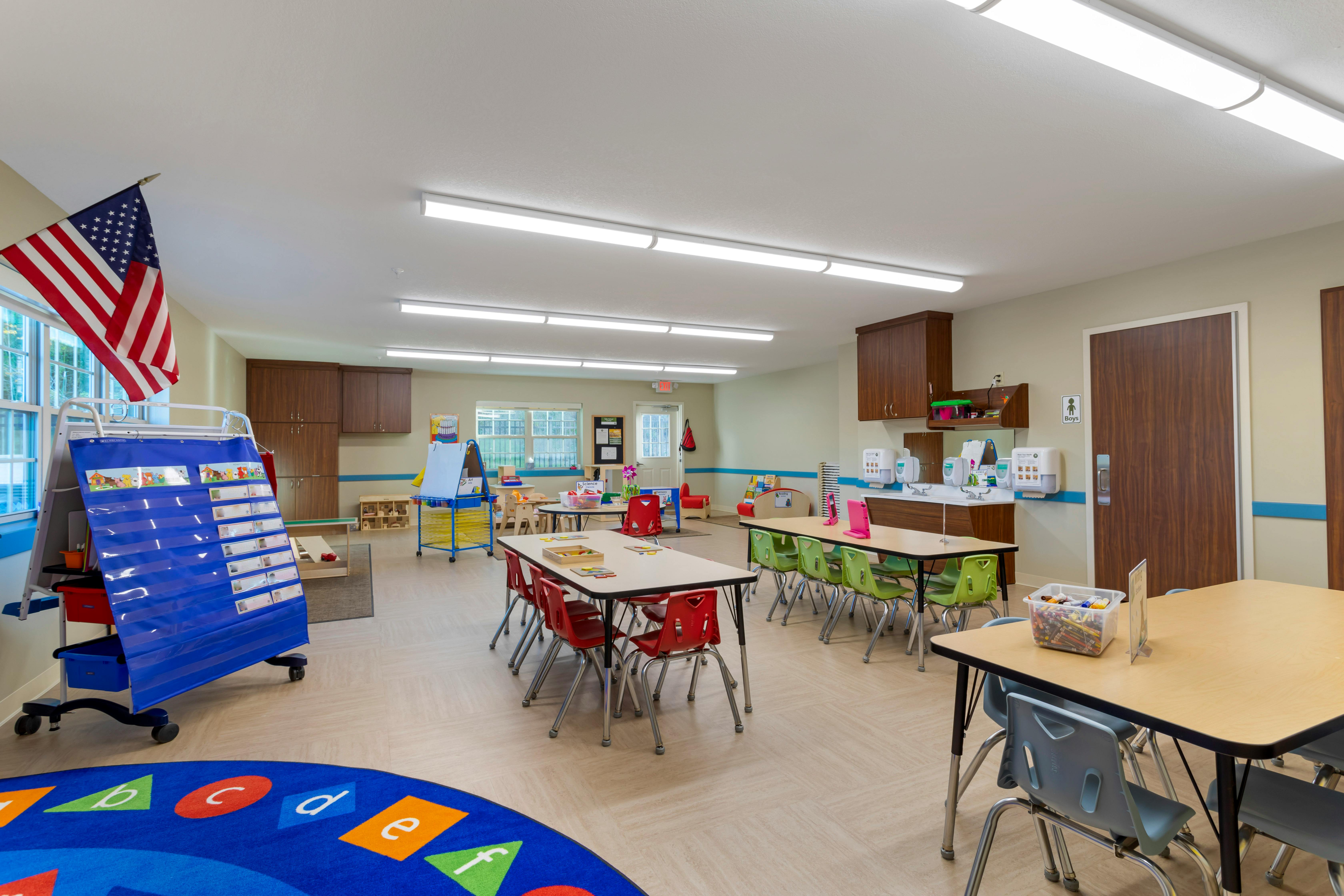 Classroom with small tables and chairs, a blue rug with letters, and an American flag