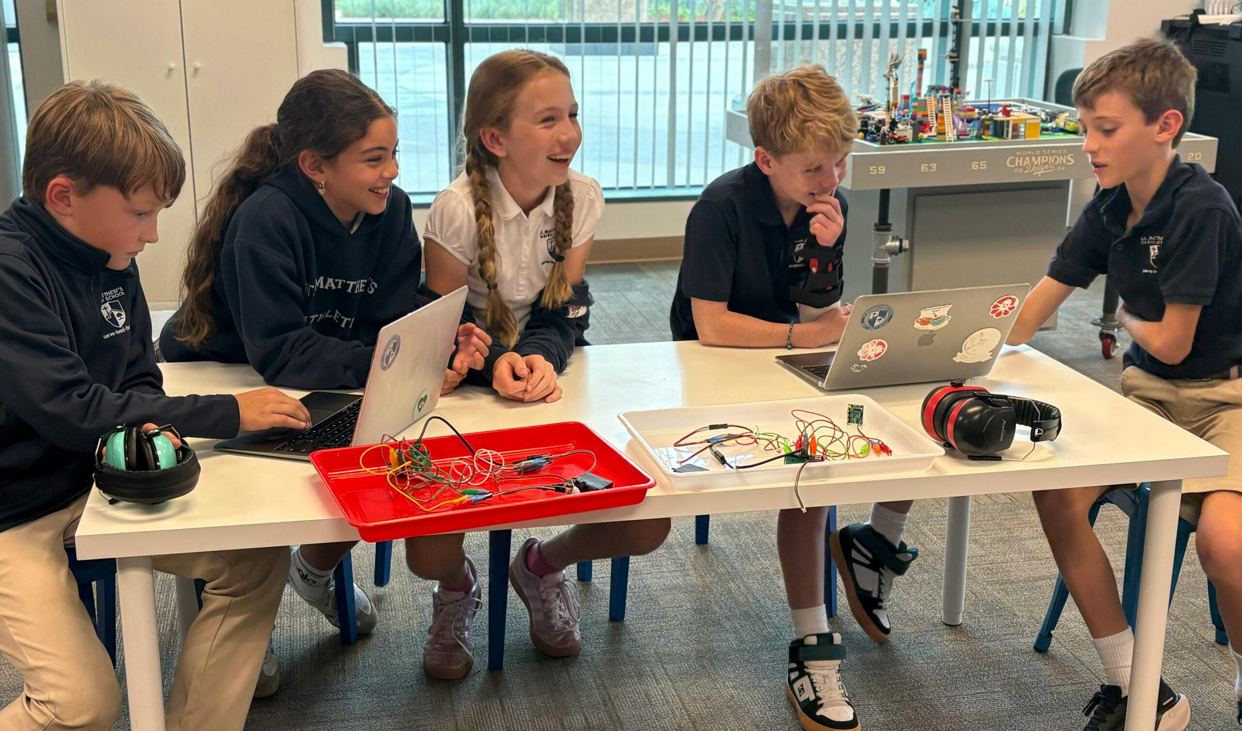Five children working on laptops and electronics at a table in a classroom