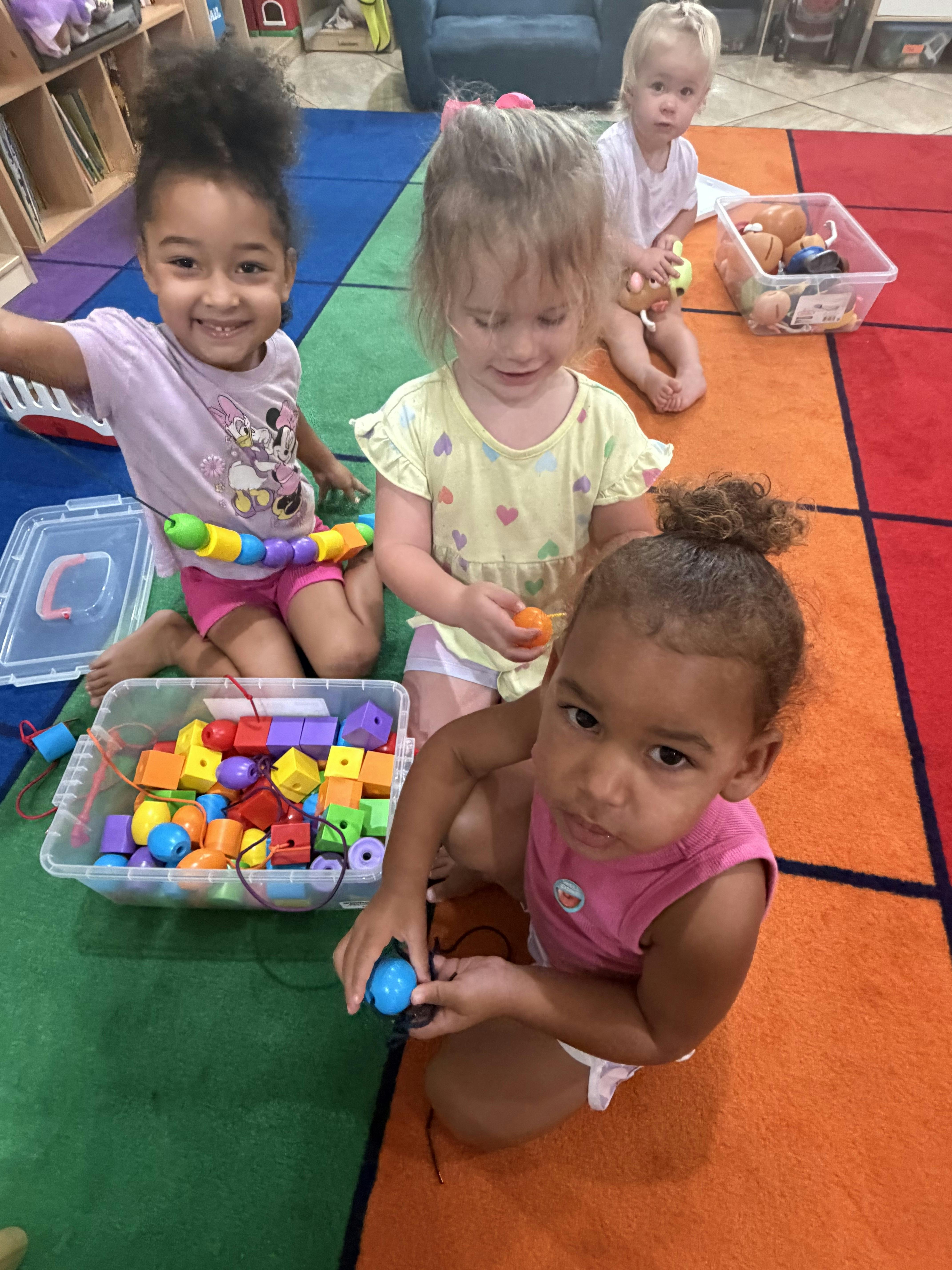 Four children sitting on a colorful classroom rug playing with plastic beads