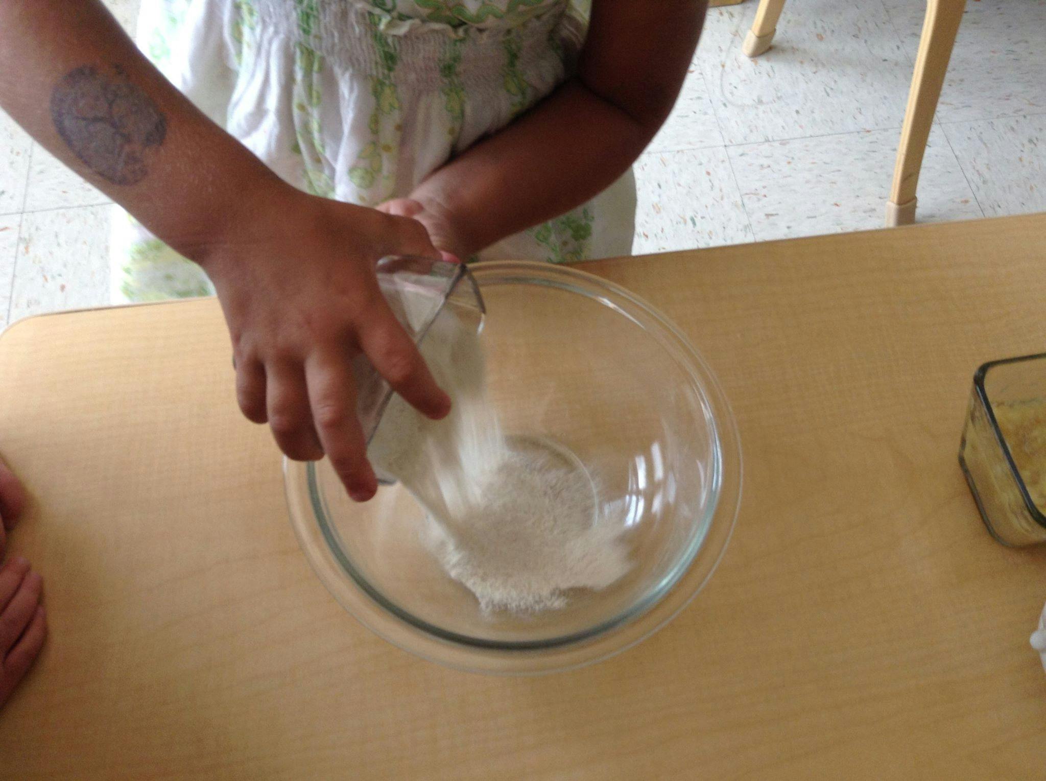 Child pouring white powder from a measuring cup into a glass bowl