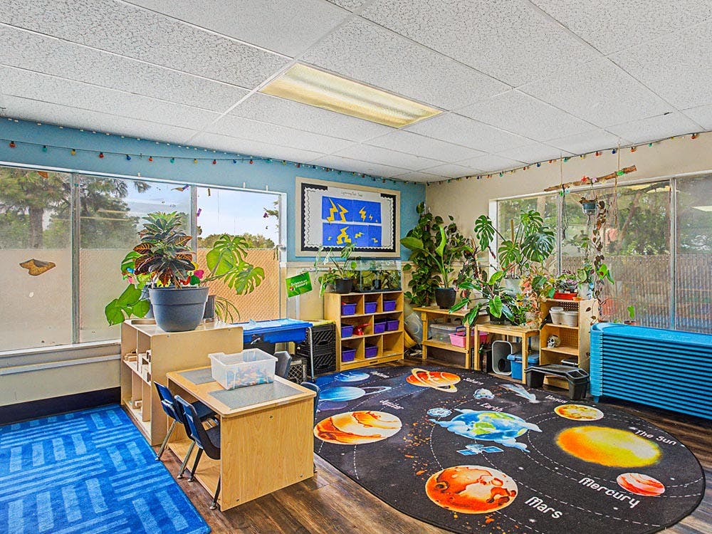 Classroom with large windows, several plants, a solar system rug, and student desks