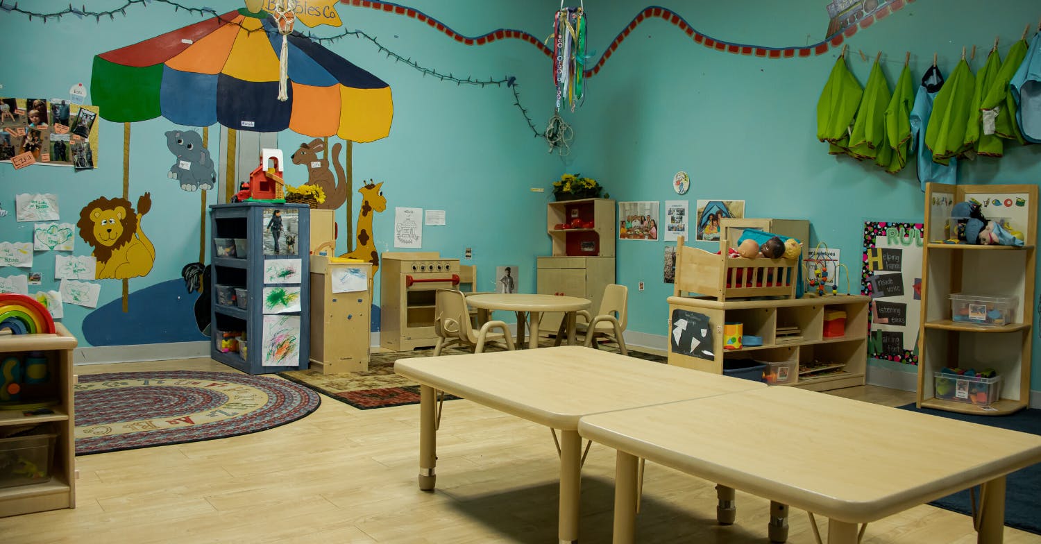 Classroom with small tables and chairs, shelves with toys, and colorful decorations